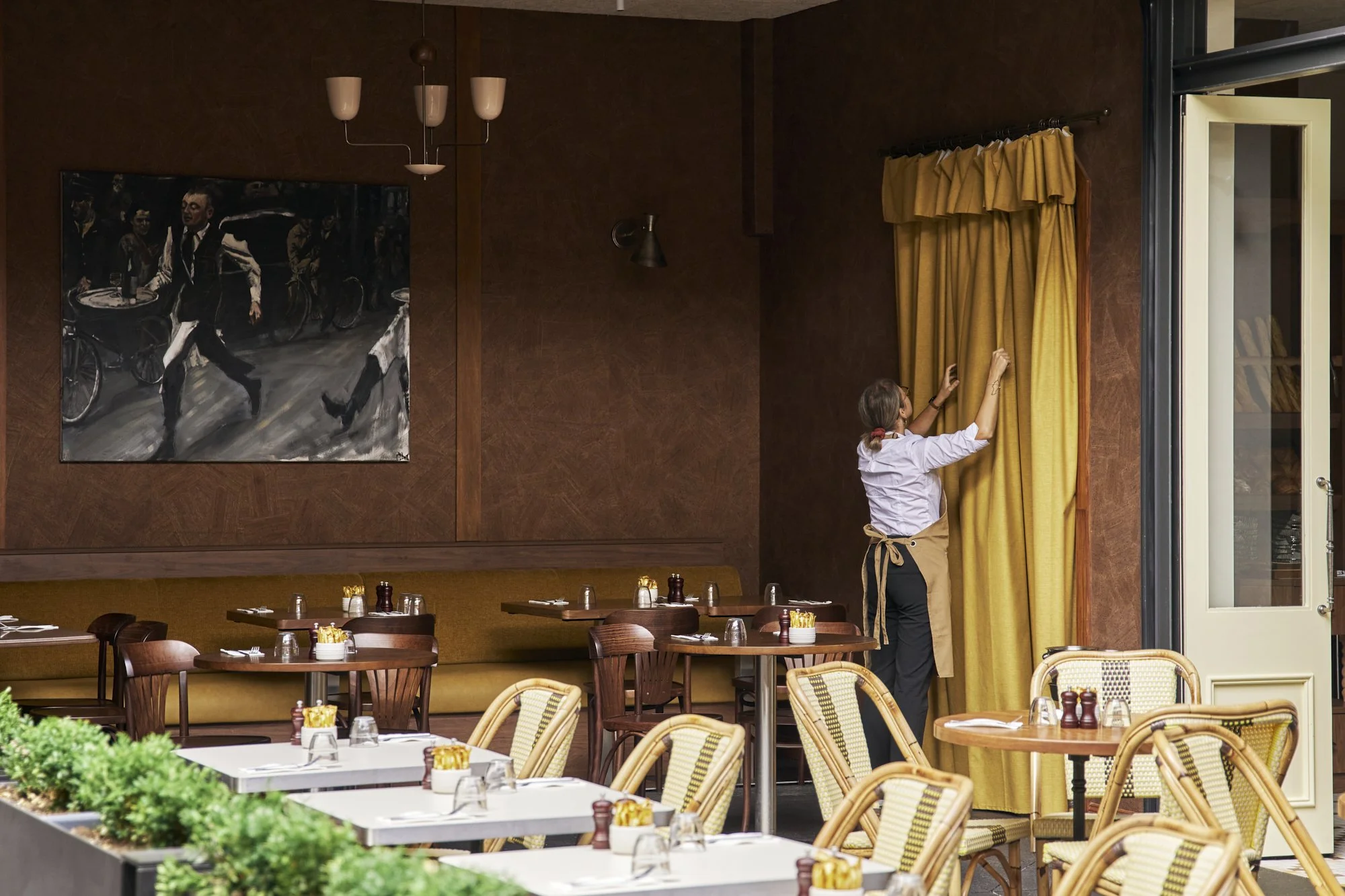 A woman in a white shirt and black pants pulls back yellow curtains in a restaurant. The restaurant has wooden walls, a large painting of people crossing a street, and tables set for dining with glasses, napkins, and condiments.