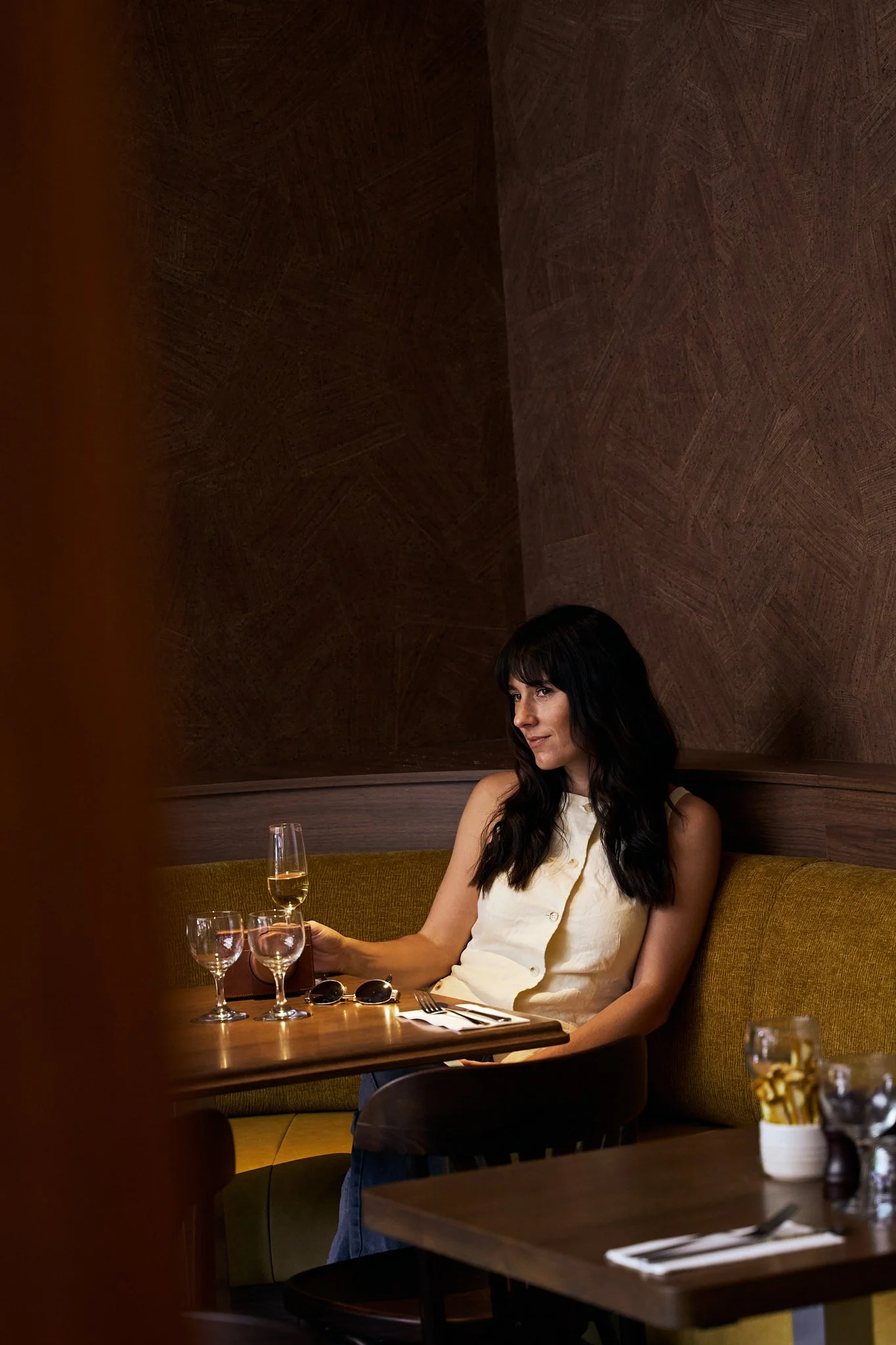 Woman with dark hair sitting alone at a table in a restaurant, holding a glass of champagne, with glasses and utensils on the table, in a dimly lit restaurant with brown walls.