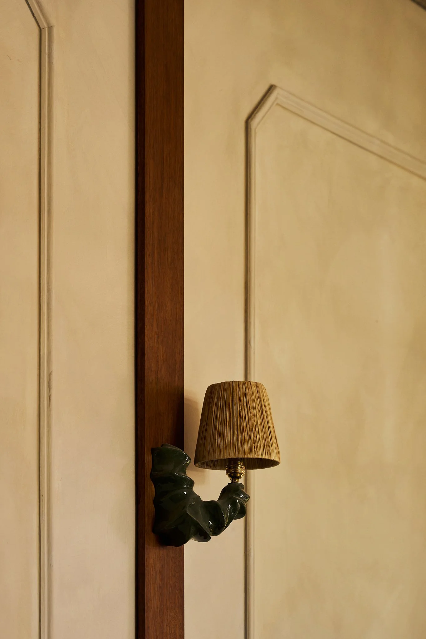 Wall-mounted lamp with a cone-shaped lampshade and a decorative base resembling a curled leaf, attached to a wooden panel on a beige wall with decorative molding.