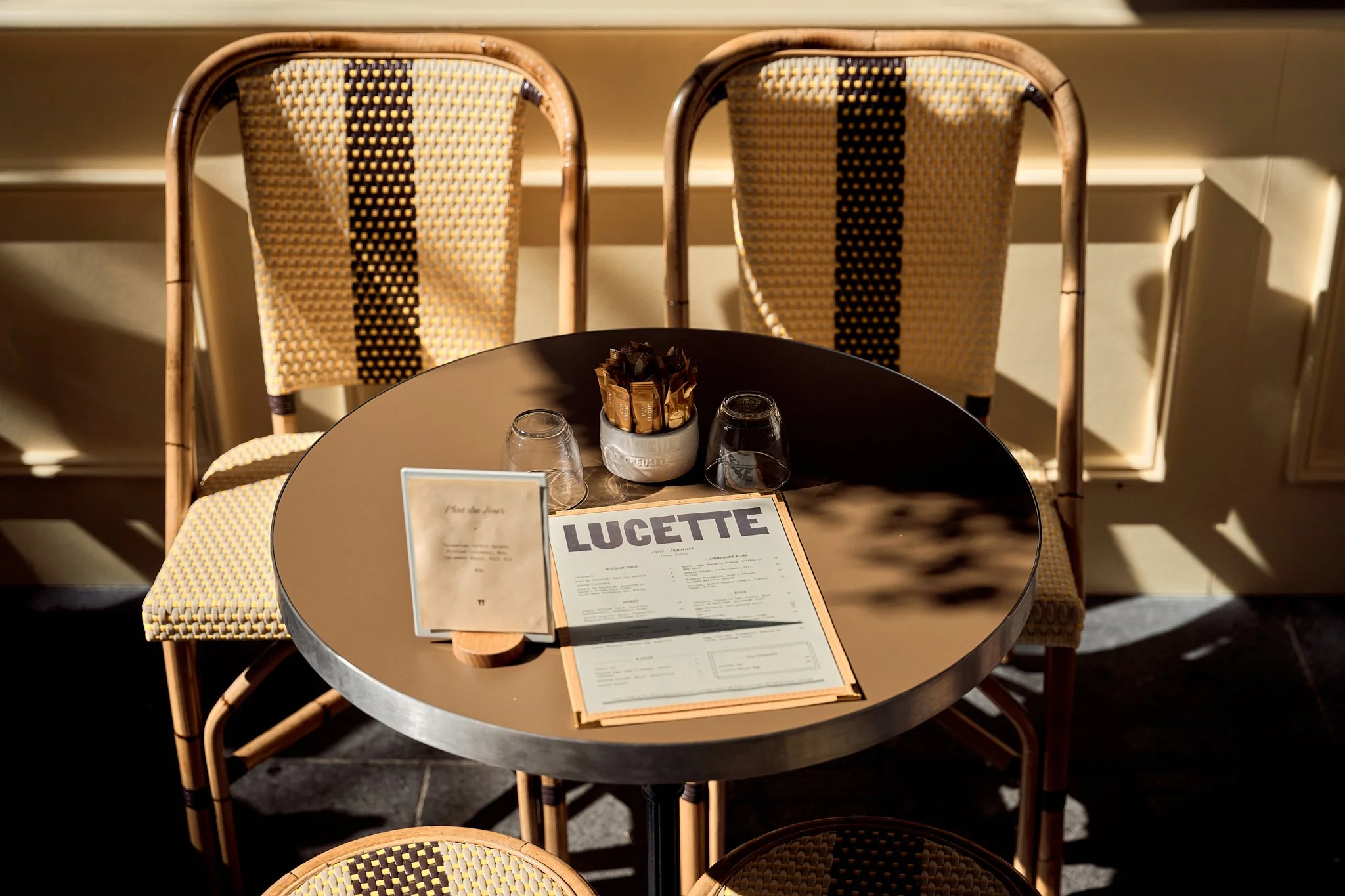 A round black table with two wicker chairs with yellow and black woven seats behind it, set in a sunny indoor or outdoor cafe. On the table are a menu, a small stand-up menu, three upside-down glasses, and a container holding sugar packets.