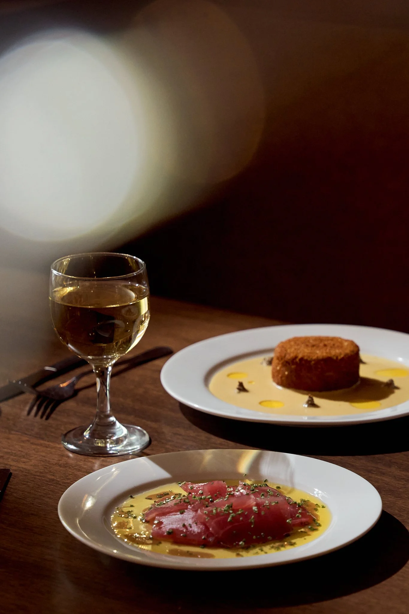 A glass of white wine, a plate with a coated appetizer, and a plate with slices of raw fish garnished with herbs on a wooden table.
