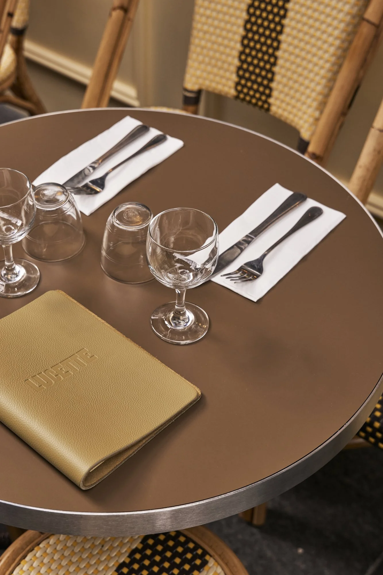 Table setting with two wine glasses, two coffee cups upside down, two sets of utensils on white napkins, a yellow menu or notebook with the word "HUE" embossed, and a wicker chair in the background.