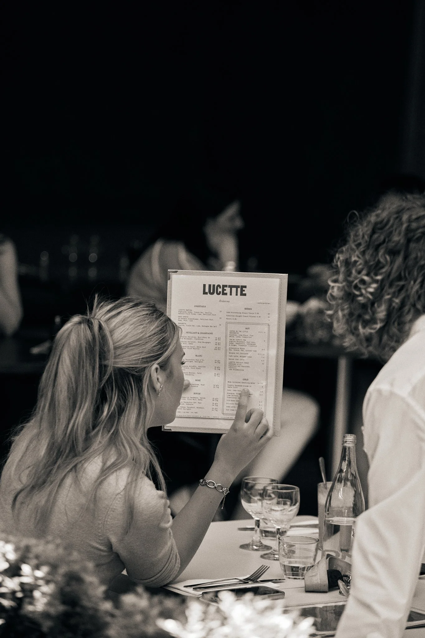 A woman at a restaurant table holding a menu titled 'LUCETTE' while looking at it, with a woman in the background and dinnerware set on the table in black and white.