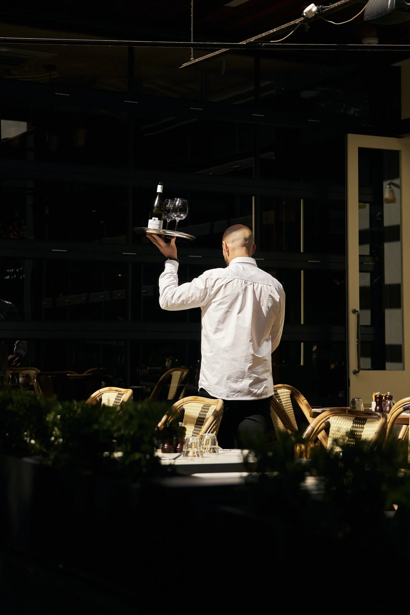 A waiter dressed in a white shirt carries a tray with empty wine glasses and a wine bottle in a dimly lit restaurant or cafe.
