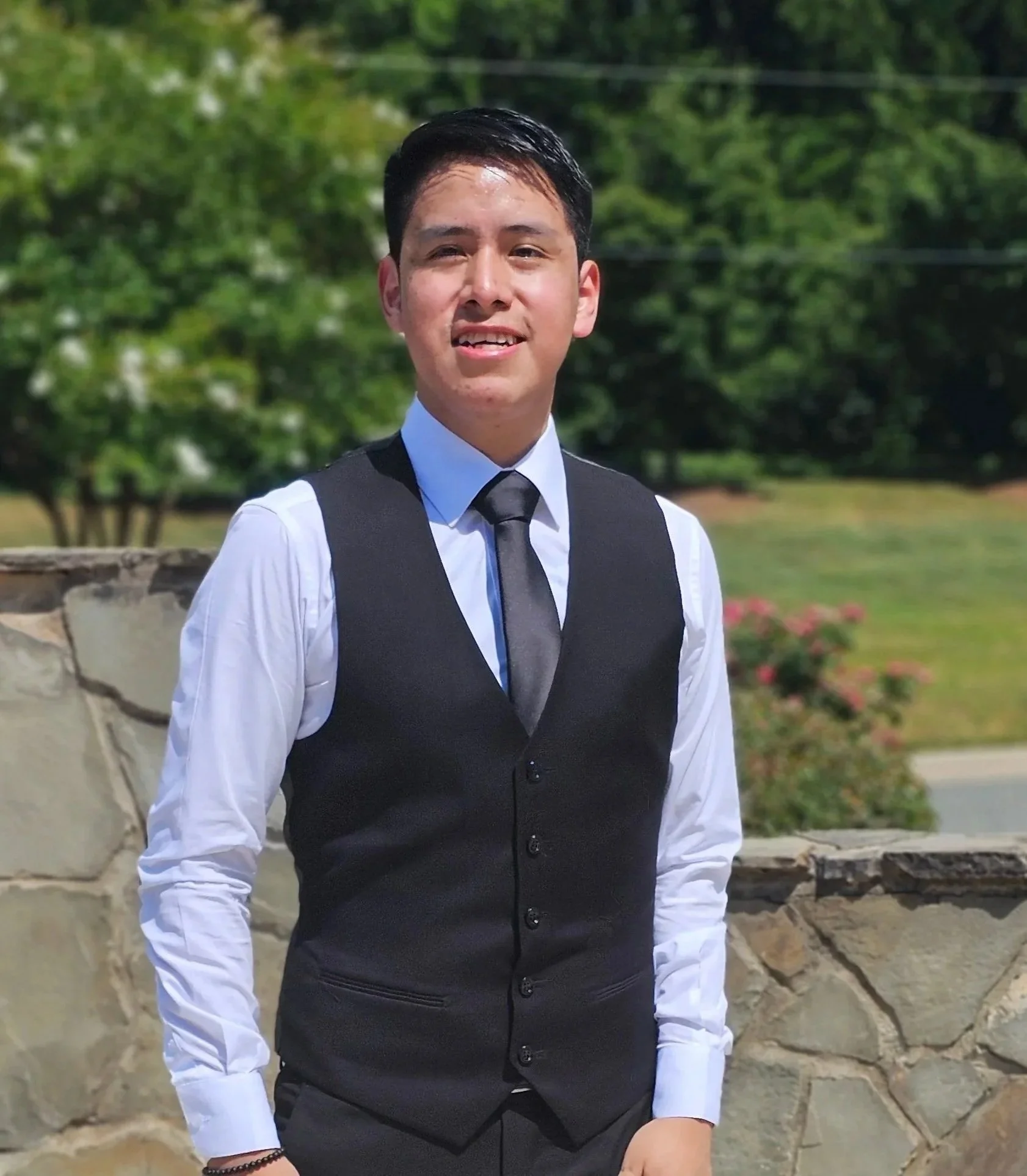 A young man wearing formal attire, including a black vest, white shirt, and black tie, standing outdoors with a stone wall and green trees in the background.