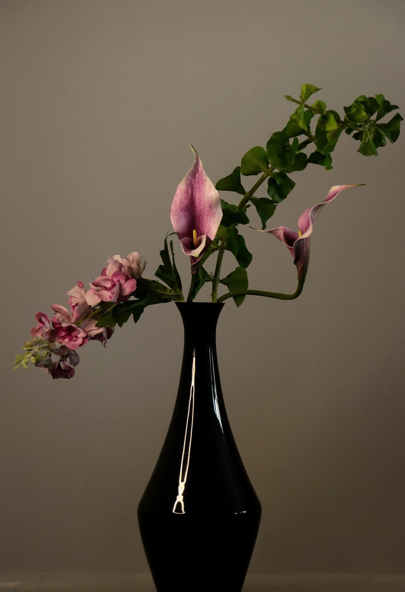 A black vase holding a floral luxury arrangement with pink and purple flowers and green curve leaves on a plain background.