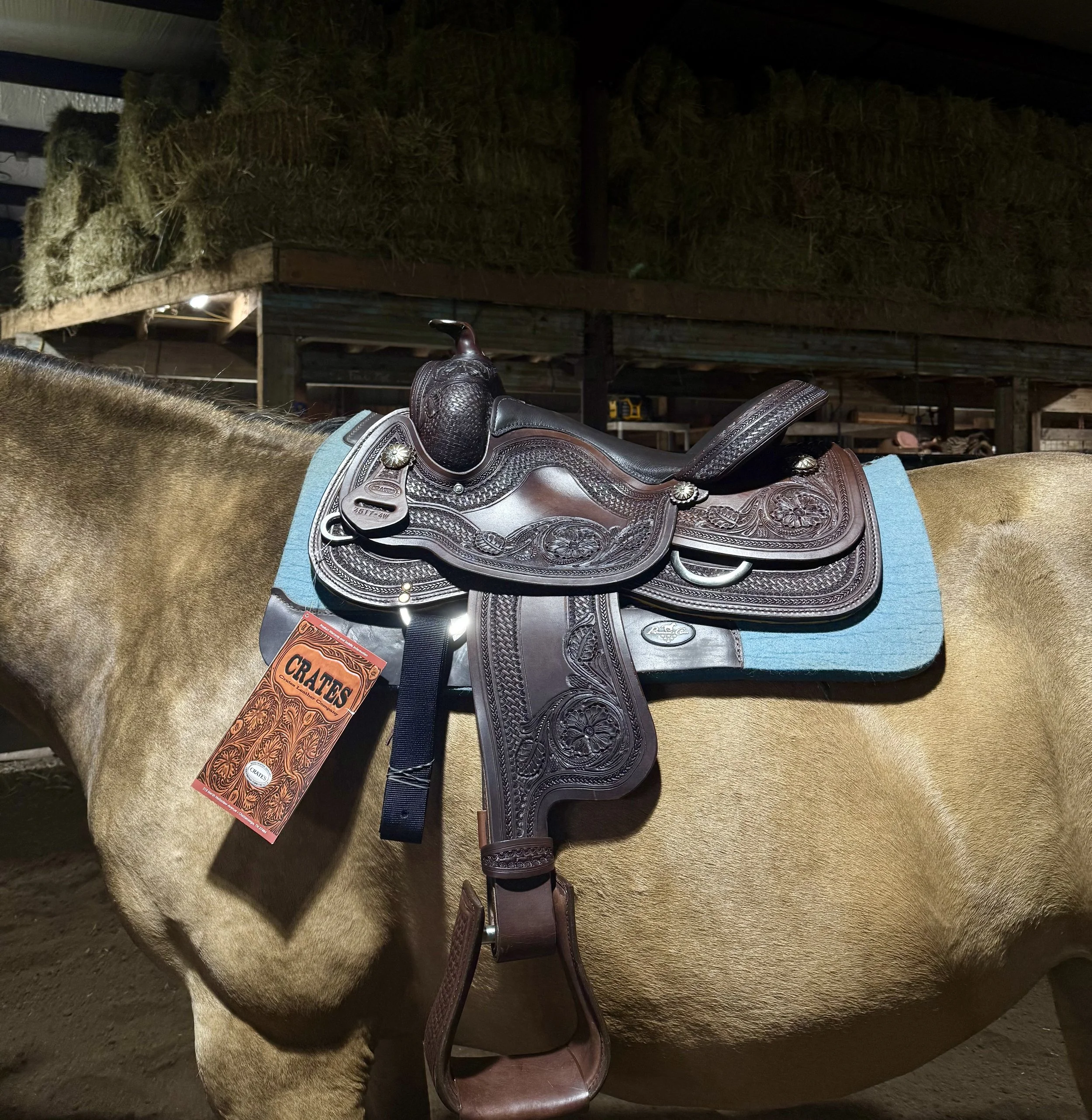 A horse with a brown saddle, stirrups, and a blue saddle pad standing next to a hay bale in a barn or stable.