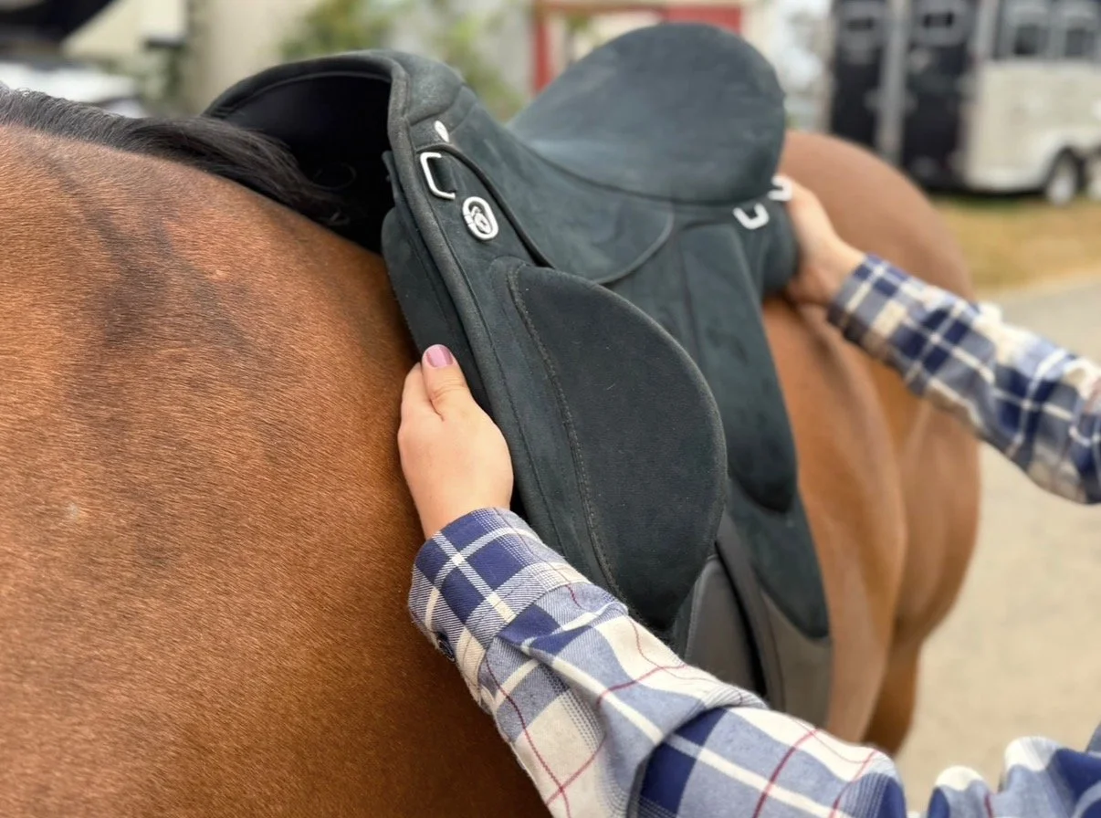 A person with a plaid shirt adjusting a saddle on a brown horse.
