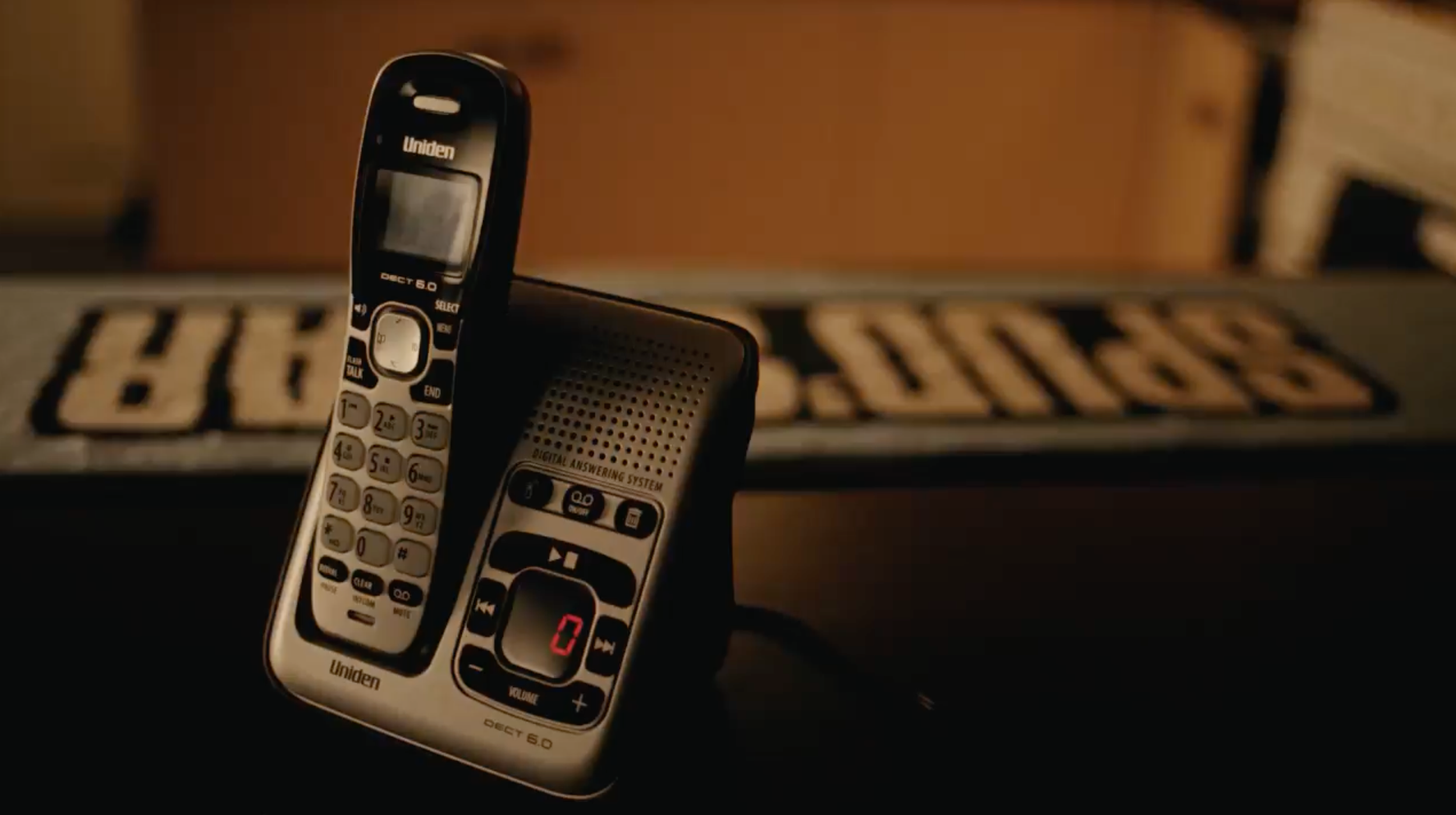 A cordless landline phone placed on a stereo speaker, with a blurred advertisement or label in the background.