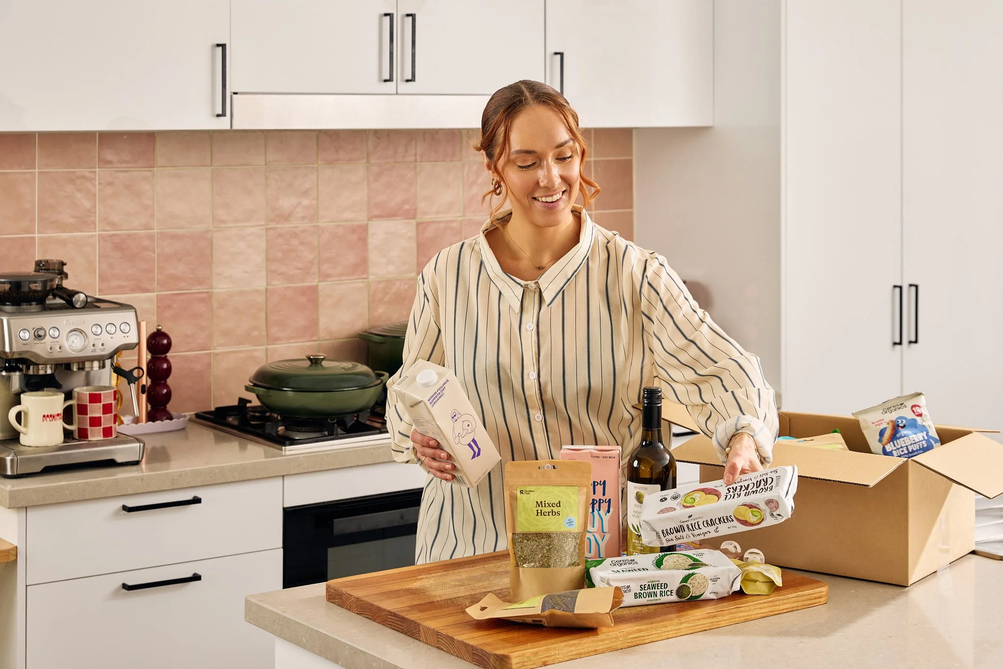 A woman with reddish hair wearing a beige striped shirt is in a kitchen. She is smiling and looking at food ingredients on a wooden cutting board on the counter. She holds a carton of soy milk in her right hand and a paper bag in her left hand. The kitchen has white cabinets, pink tiled backsplash, and various cooking items including a coffee machine, pots, and food packages on the countertop and in a cardboard box.