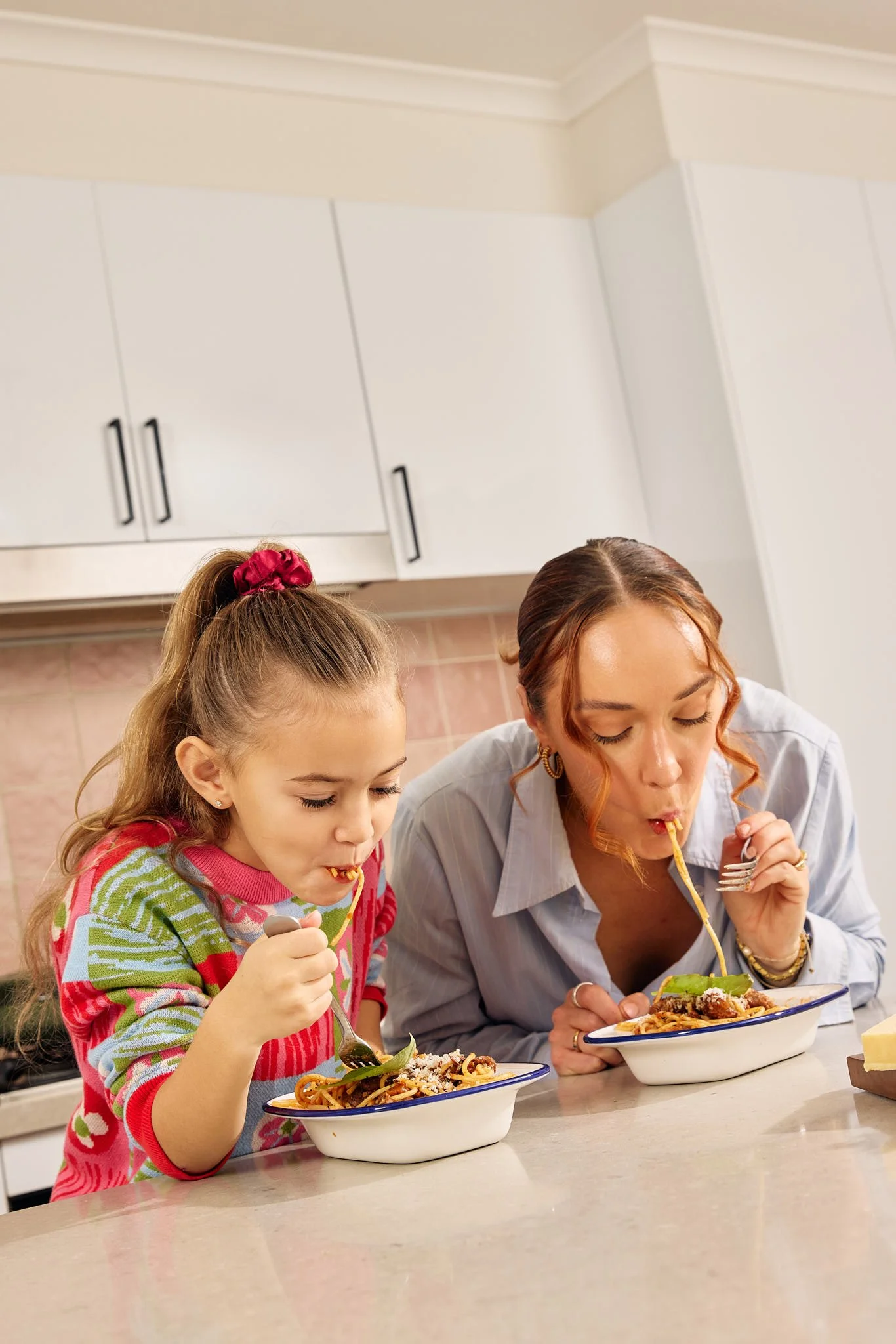 A woman and a young girl are eating spaghetti with salad in a kitchen.