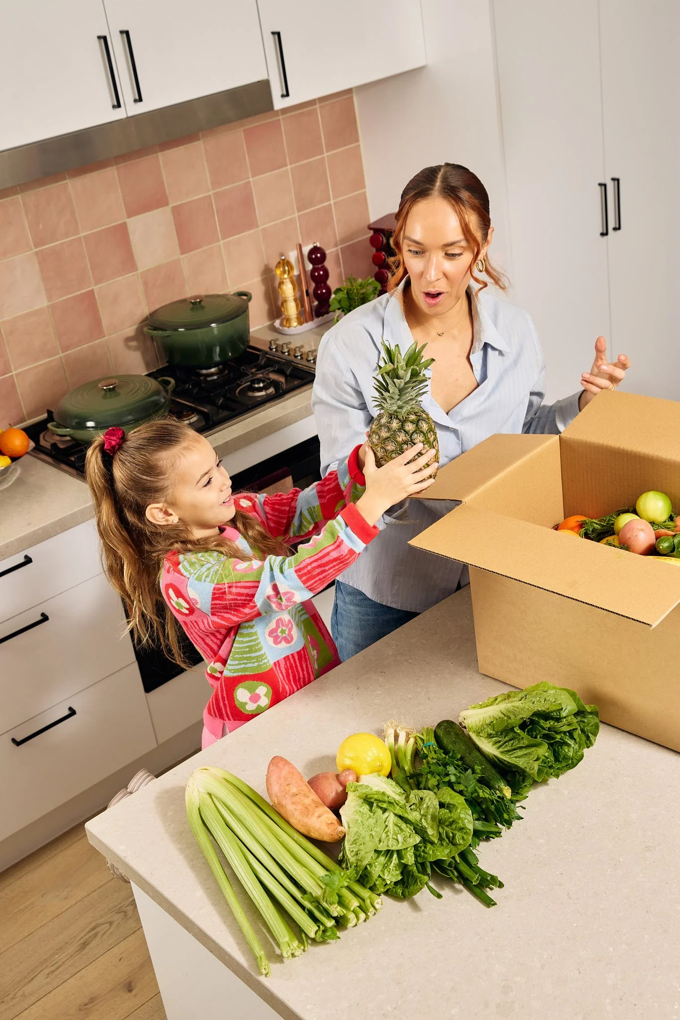 A woman and a girl are unpacking groceries in a kitchen. The woman holds a pineapple, and the girl is reaching for it. On the counter, there are assorted vegetables including lettuce, celery, sweet potatoes, and other produce, with a large cardboard box containing fruits and vegetables.