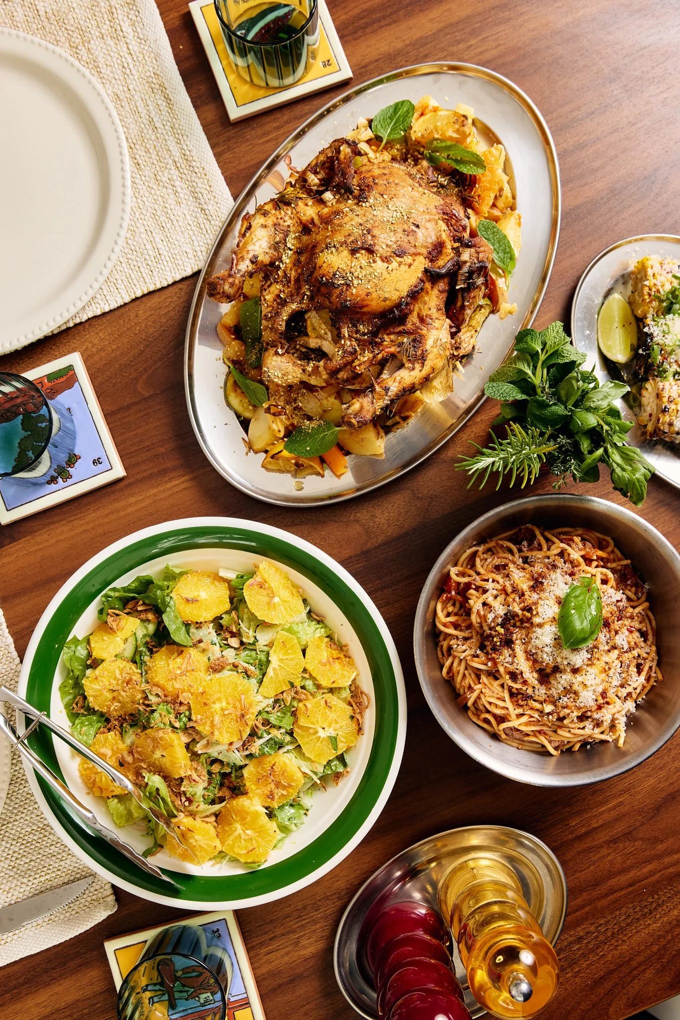 A table set with various dishes including a roasted chicken with vegetables, a green salad with orange slices, spaghetti topped with cheese, and a plate of rice and vegetables, along with a glass of white wine, a glass of red wine, and a shot glass with a yellow drink.