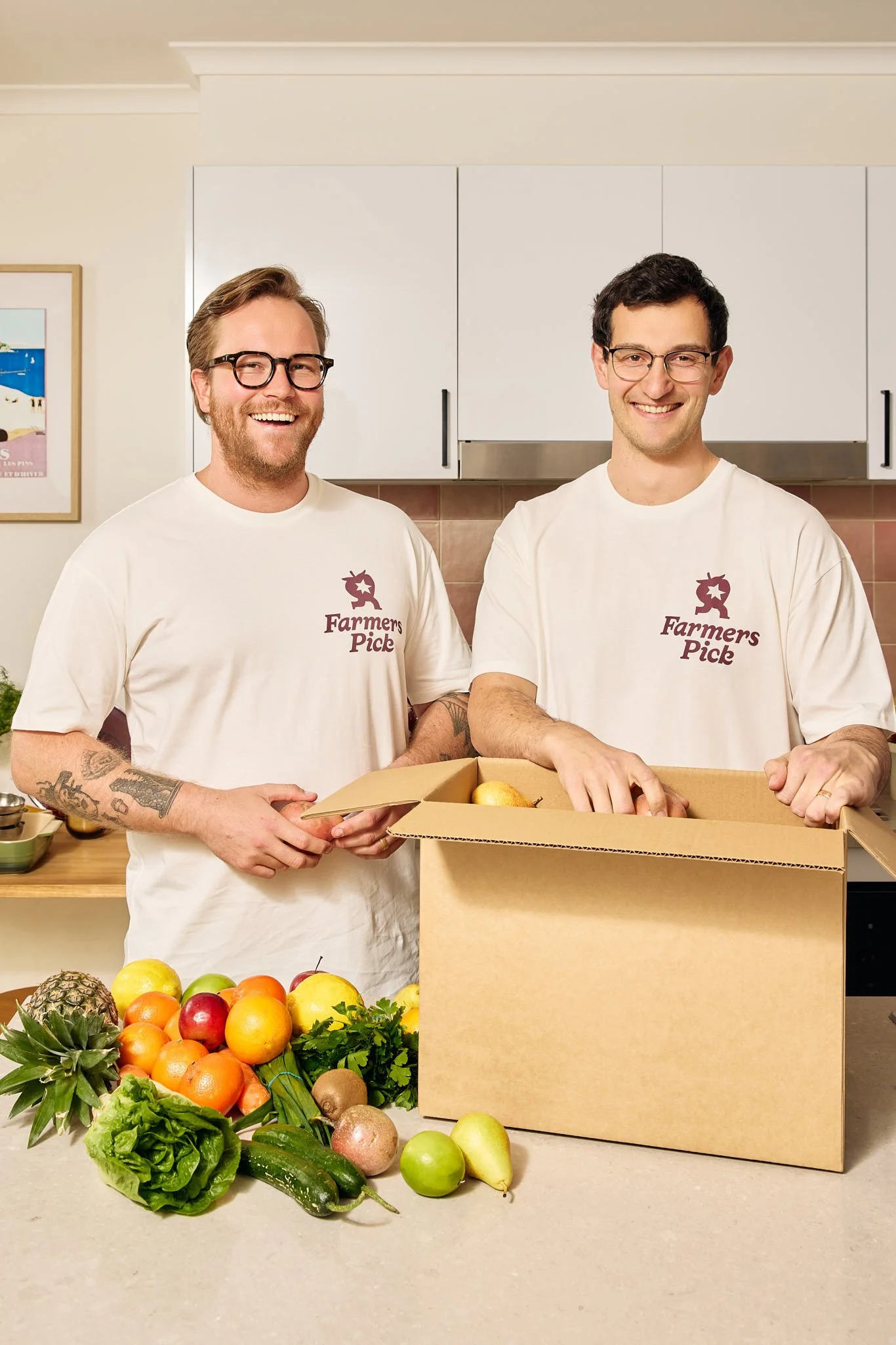 Two men wearing white shirts with "Farmers Pick" logo, smiling in a kitchen, holding a cardboard box with fruit, with fresh fruits and vegetables on a counter in front.