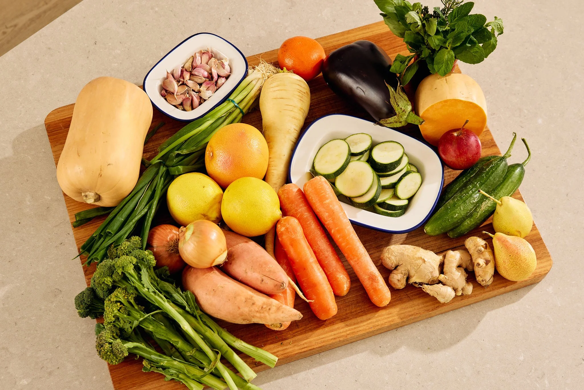 Fresh vegetables on a wooden cutting board, including garlic, green beans, ginger, carrots, lemons, apples, cucumber, eggplant, and leafy greens.