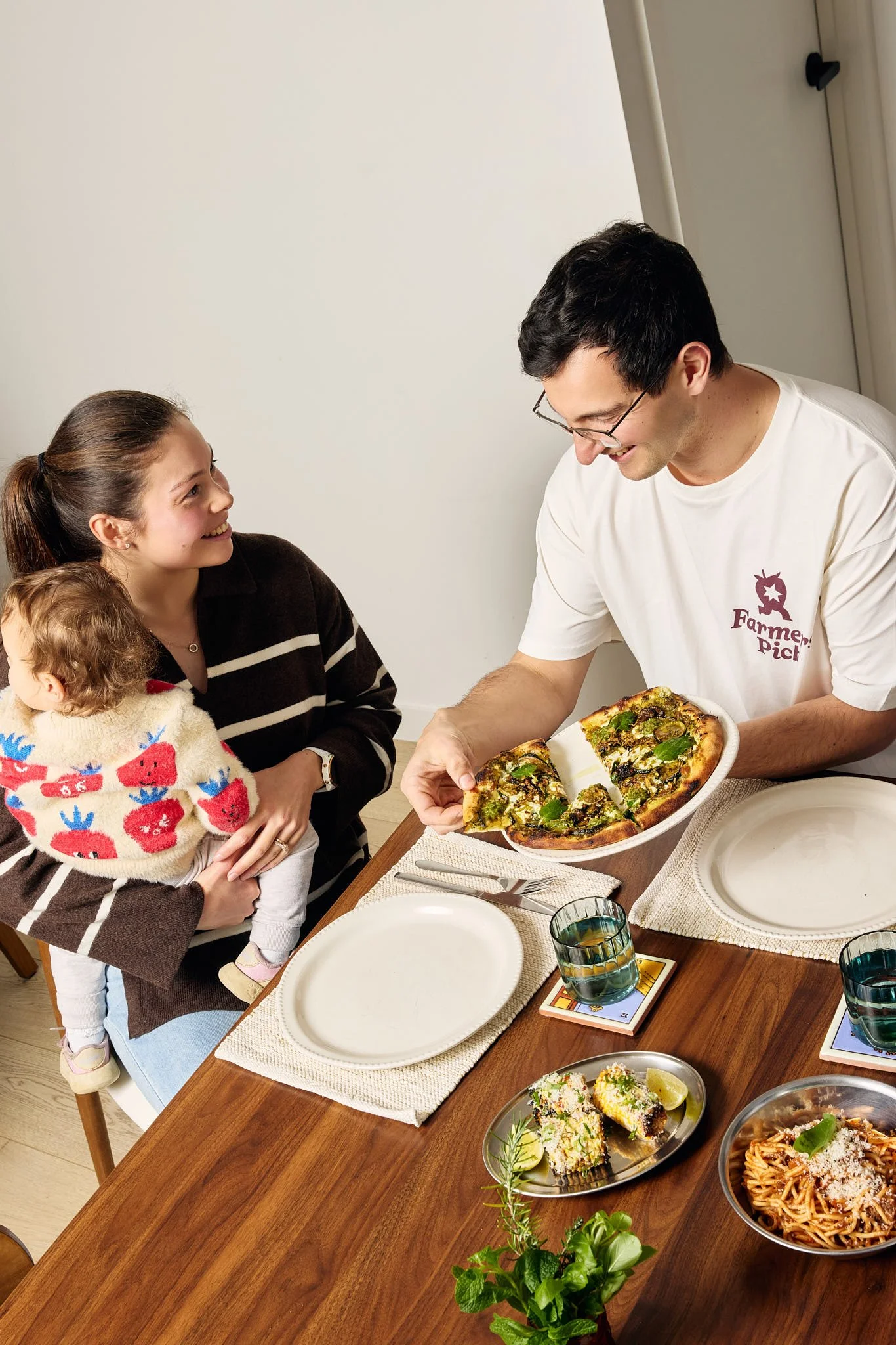 A family gathering at a dining table with friends celebrating a meal. An adult male is serving pizza to a young woman holding a toddler, with various dishes and drinks on the table.
