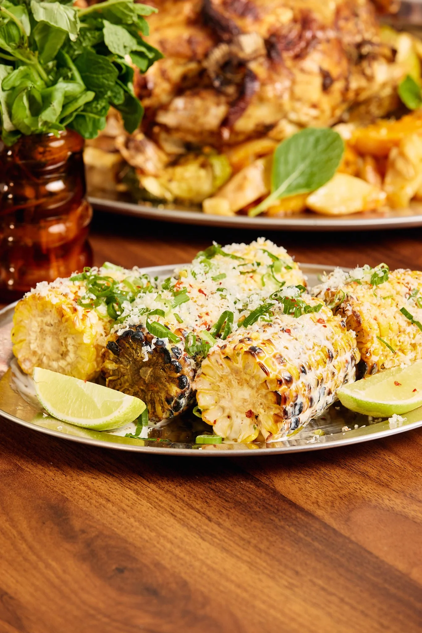 Grilled corn on the cob garnished with chopped green onions and lime wedges on a silver platter.