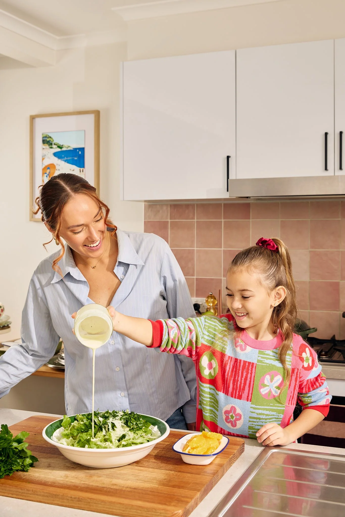 A woman and a girl are preparing a salad together in a kitchen. The woman is pouring dressing onto a large bowl of salad, and the girl is holding a bottle of dressing, smiling. The kitchen has white cabinets and a colorful picture on the wall.
