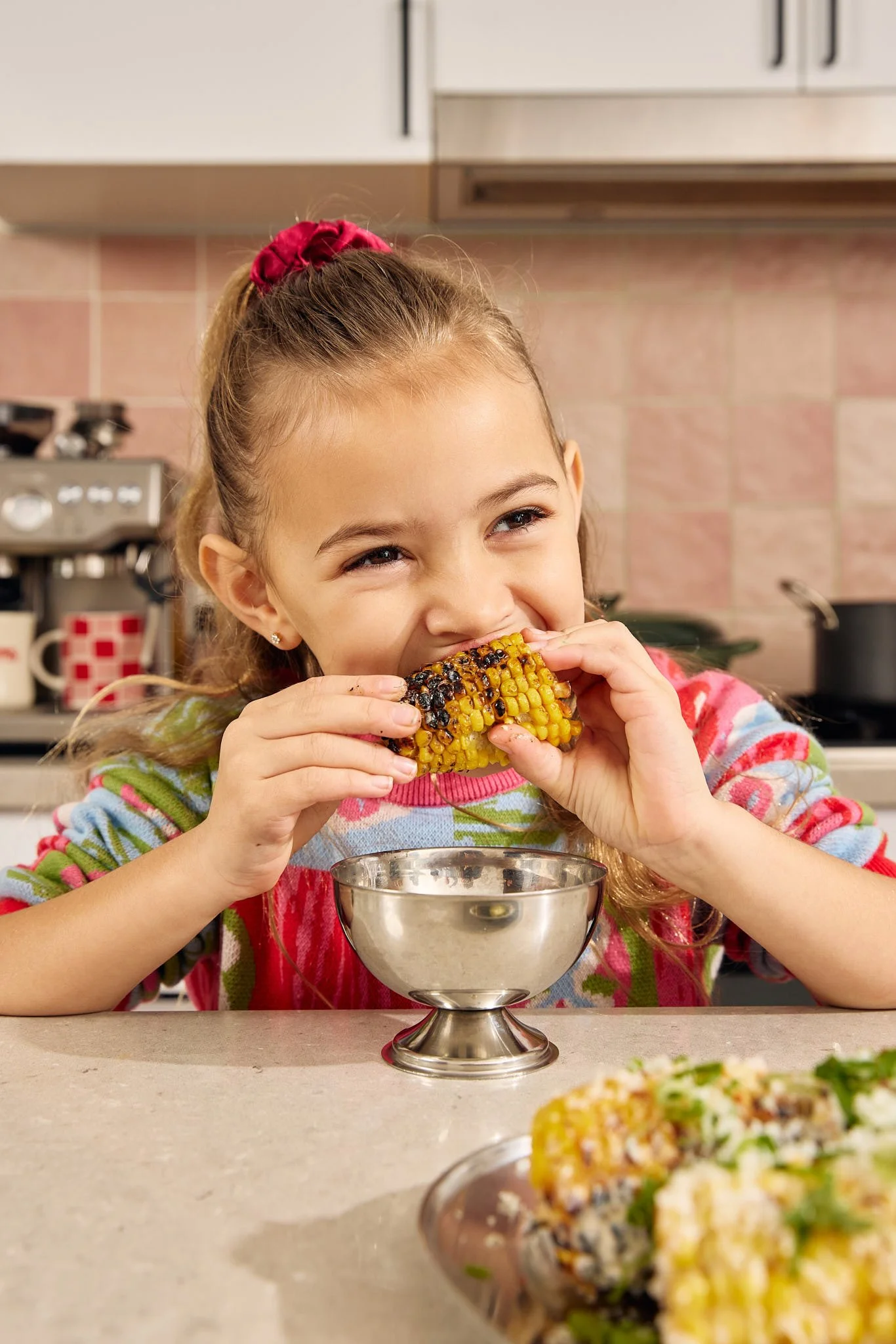 A young girl with long hair tied back with a red scrunchie, wearing a colorful striped sweater, is sitting at a kitchen counter, smiling, and eating a grilled corn on the cob. There is a metal bowl in front of her and a plate with more food in the foreground. The background features kitchen cabinets, a stove, and tiled walls.