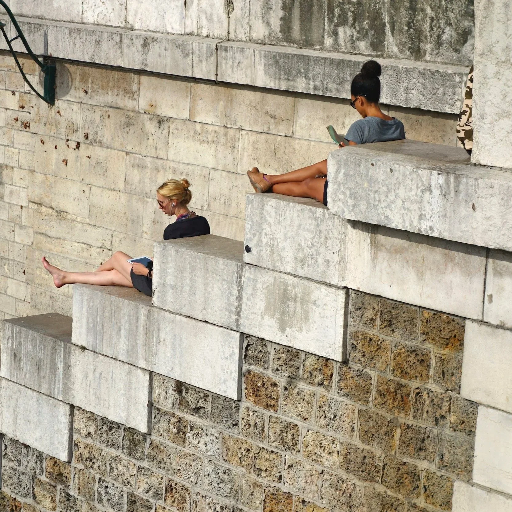 Two women and one girl sitting on large stone steps, all engaged with electronic devices. One woman is sitting on a lower step, browsing a tablet, with blond hair tied back, wearing glasses and a black outfit. The girl is sitting on an upper step, with legs extended, using a smartphone, dressed in a gray T-shirt and dark shorts. The woman in the background is sitting on an elevated step, also using a smartphone, with her hair in a bun, wearing glasses and casual clothing.