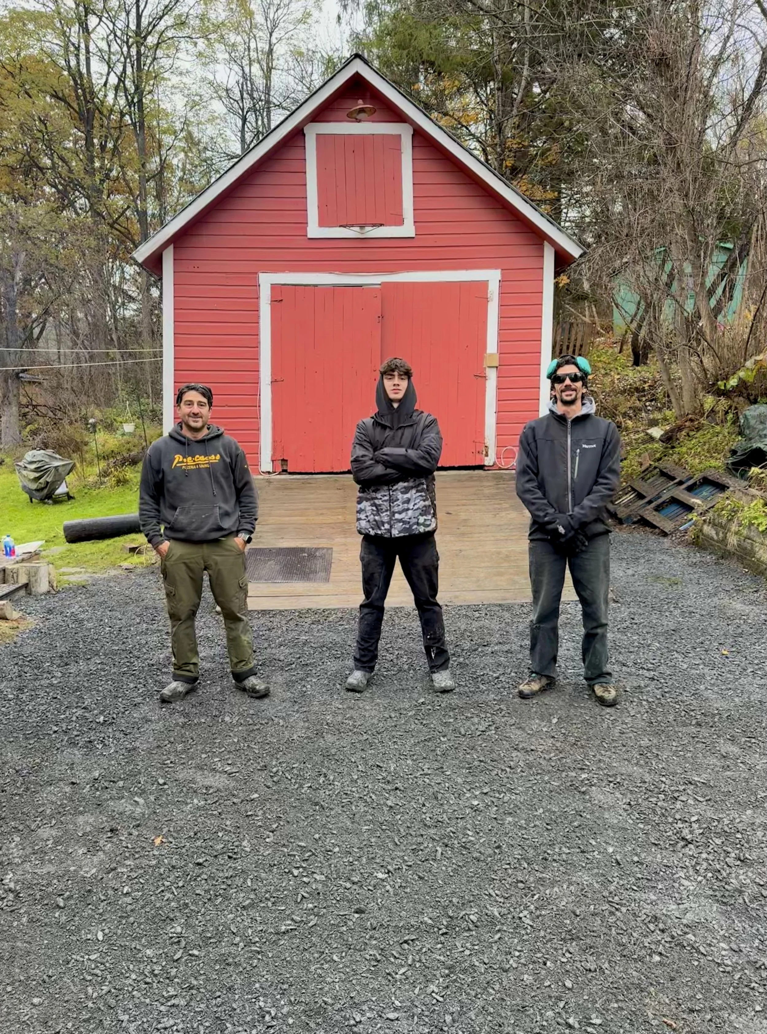 Three men standing in front of a red wooden shed with white trim, outdoors on a gravel surface, with trees in the background.