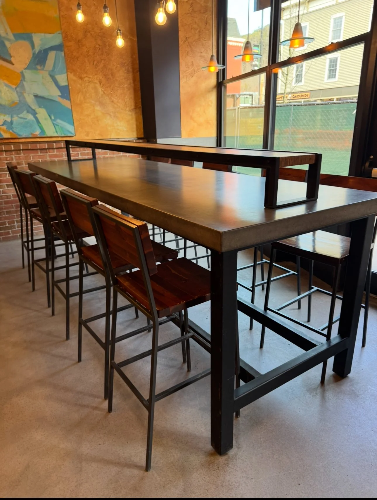 Interior of a modern restaurant with a high-top table and barstools, large windows, hanging pendant lights, and abstract mural art.