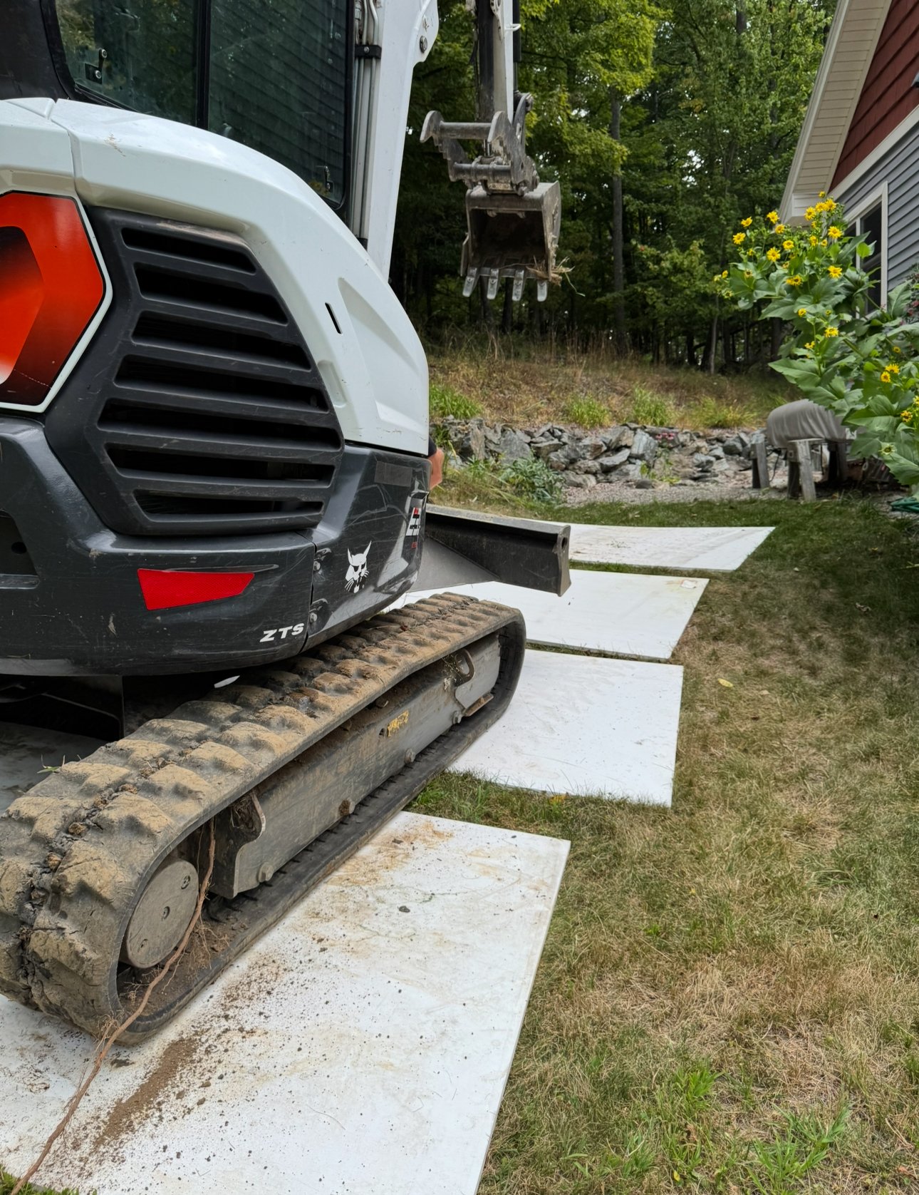 A mini excavator with tracks on concrete pads in a backyard, with a house, trees, and flowers in the background.