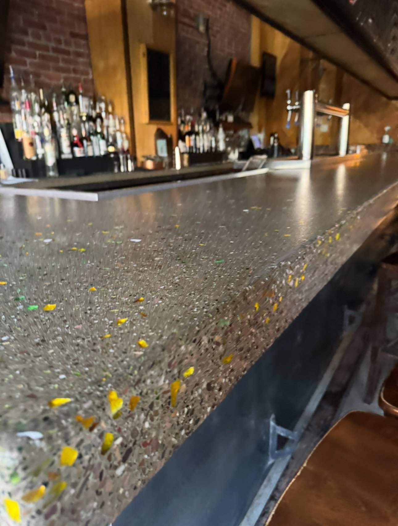 Close-up of a polished, terrazzo-style bar top with small colored chips, in a dimly lit rustic bar with bottles behind the counter.