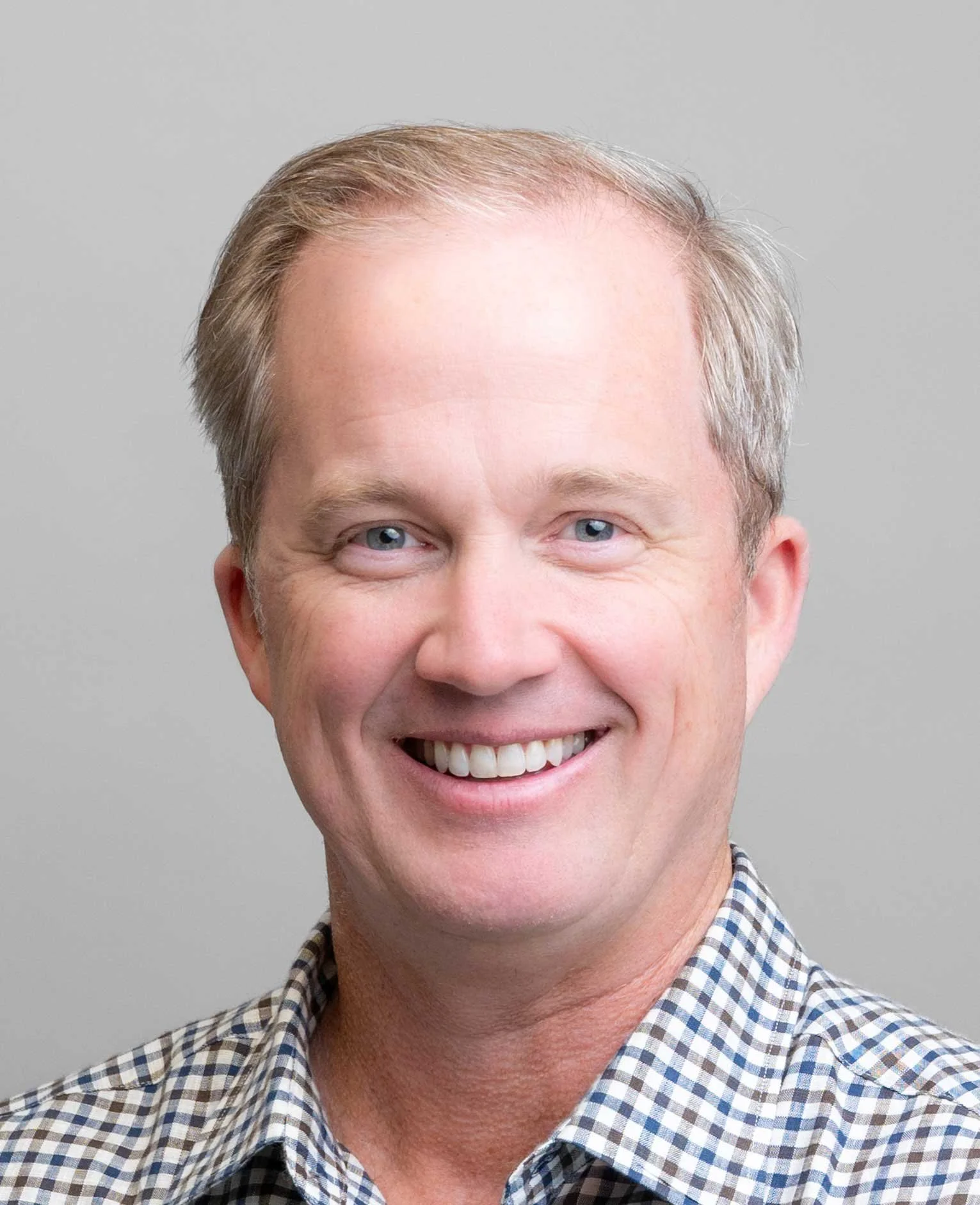 Close-up of a middle-aged man with light-colored hair, blue eyes, smiling, wearing a checkered shirt.