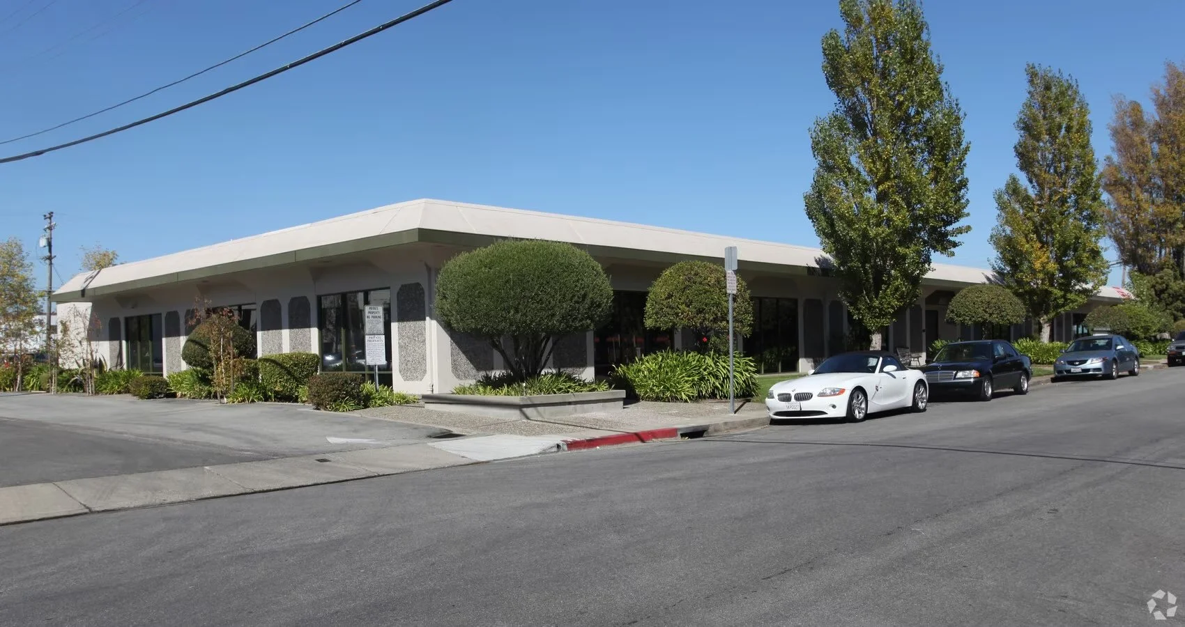 A single-story commercial building with large glass windows, surrounded by trees and shrubs, parked cars in front, and clear blue sky.