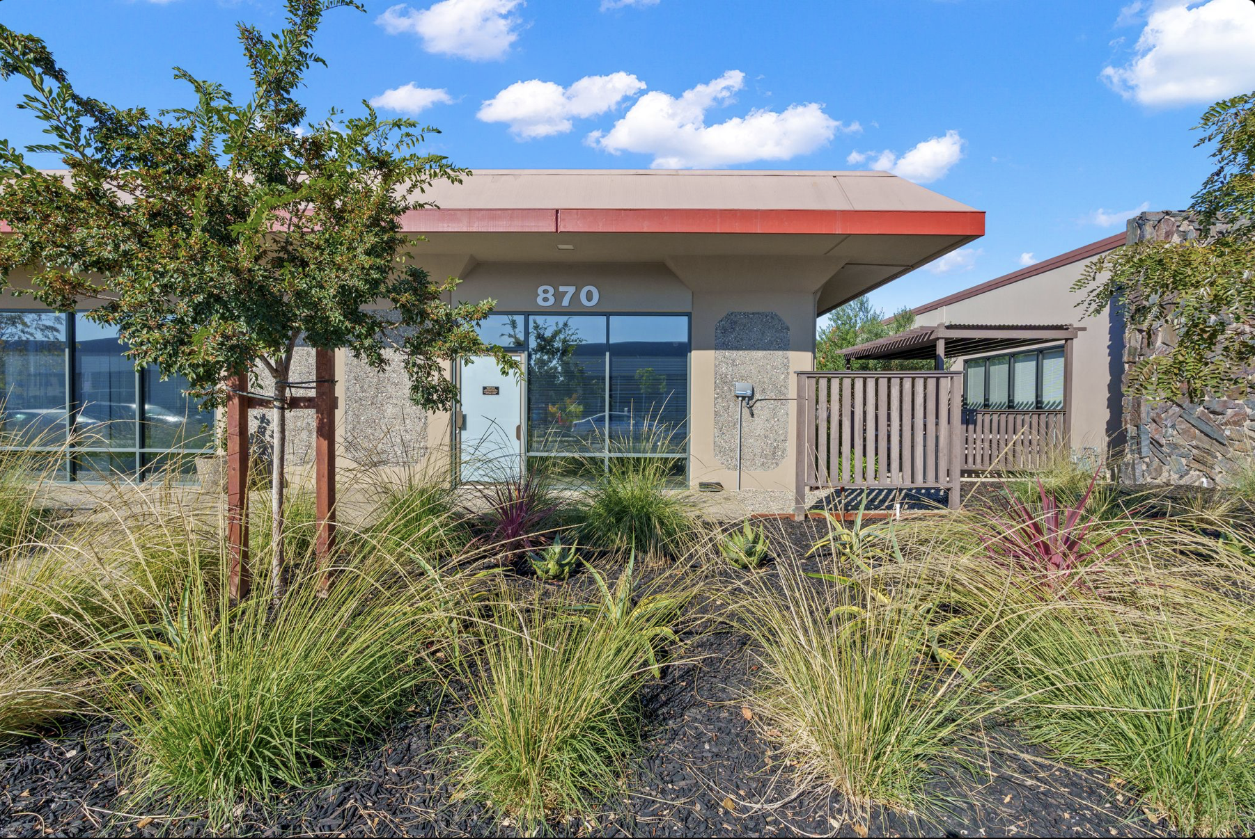 Commercial building with large glass windows, street number 870, surrounded by landscaped plants and a small tree, under a bright blue sky with scattered clouds.