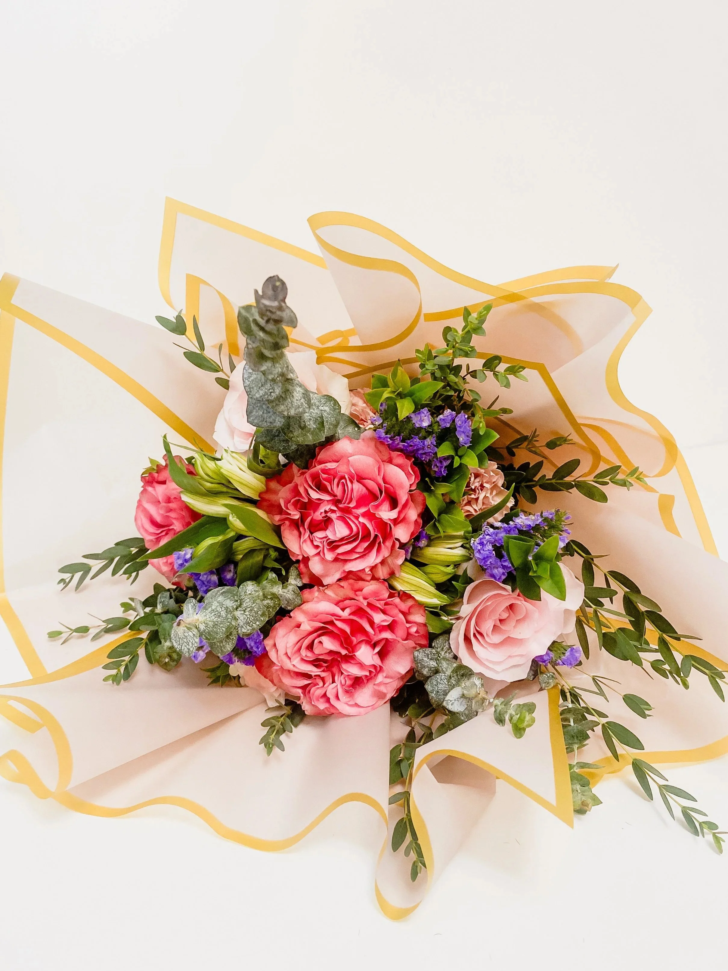 A floral bouquet featuring pink and purple flowers with green foliage, wrapped in light pink paper with yellow border accents.