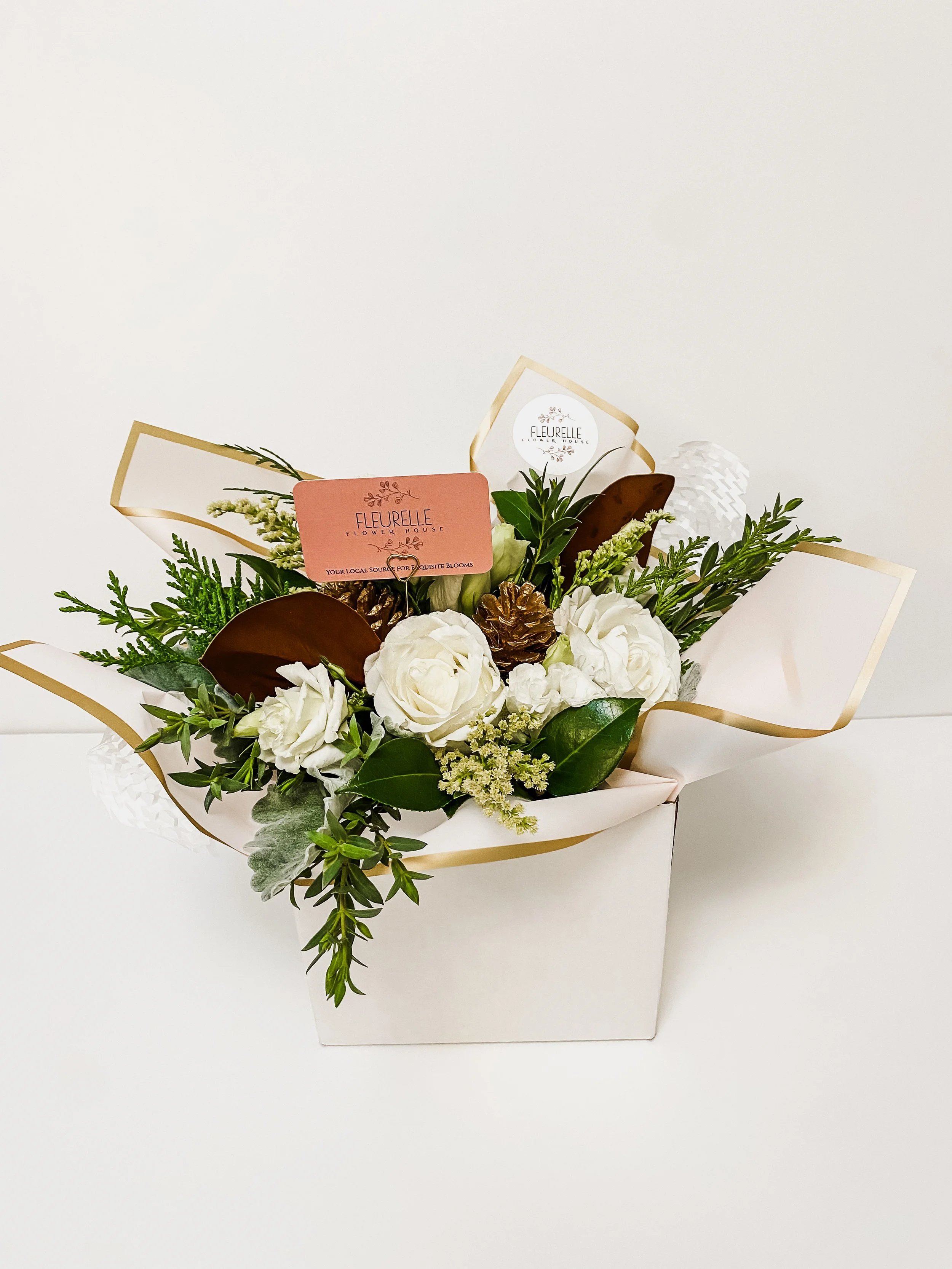 White floral arrangement in a white box with greenery, pinecones, and decorative tags from Fleurelle Flower House