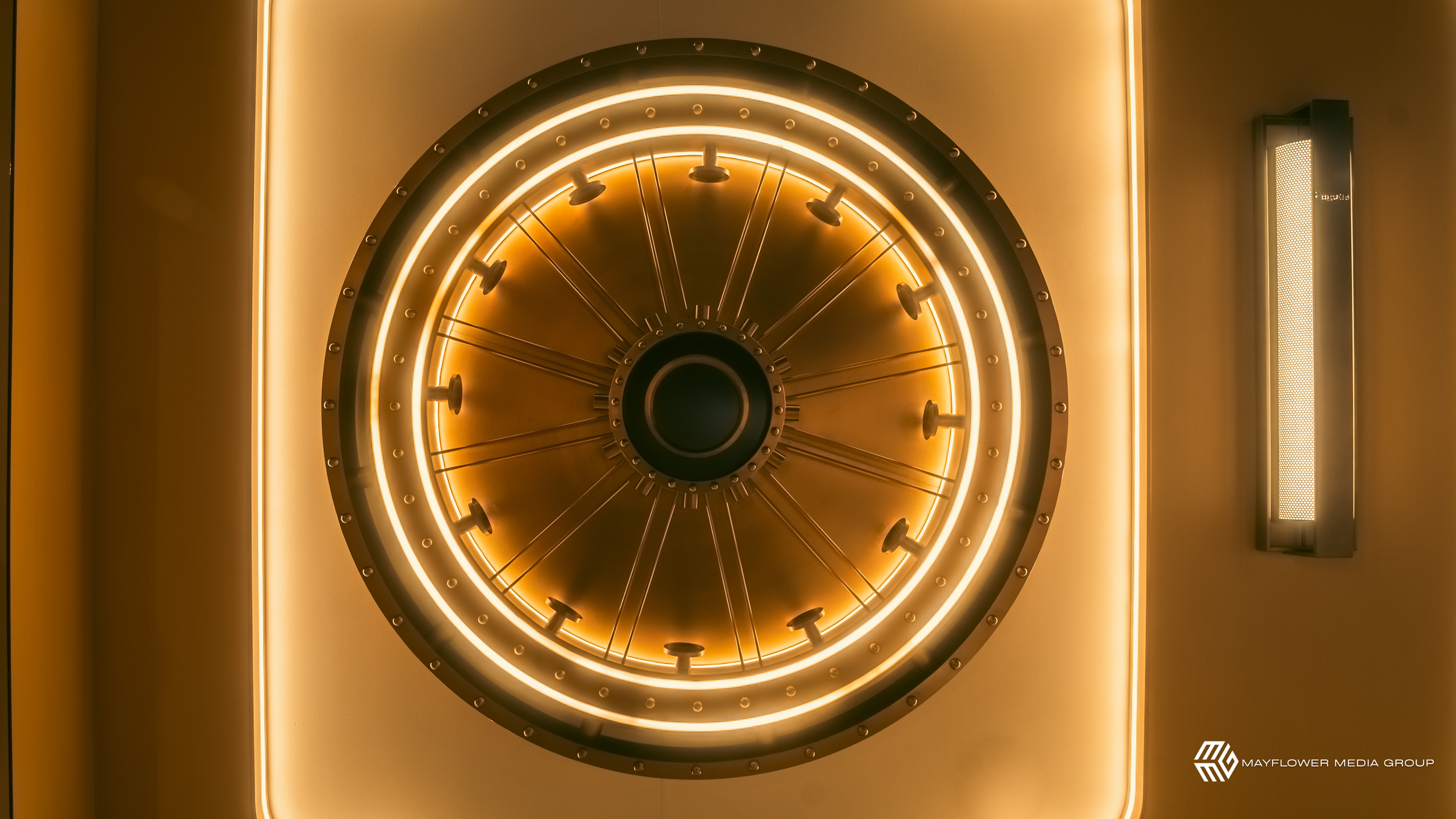 A circular, illuminated, metallic mechanism or sculpture with numerous small lights and radial spokes, mounted on a wall, with a yellowish glow.