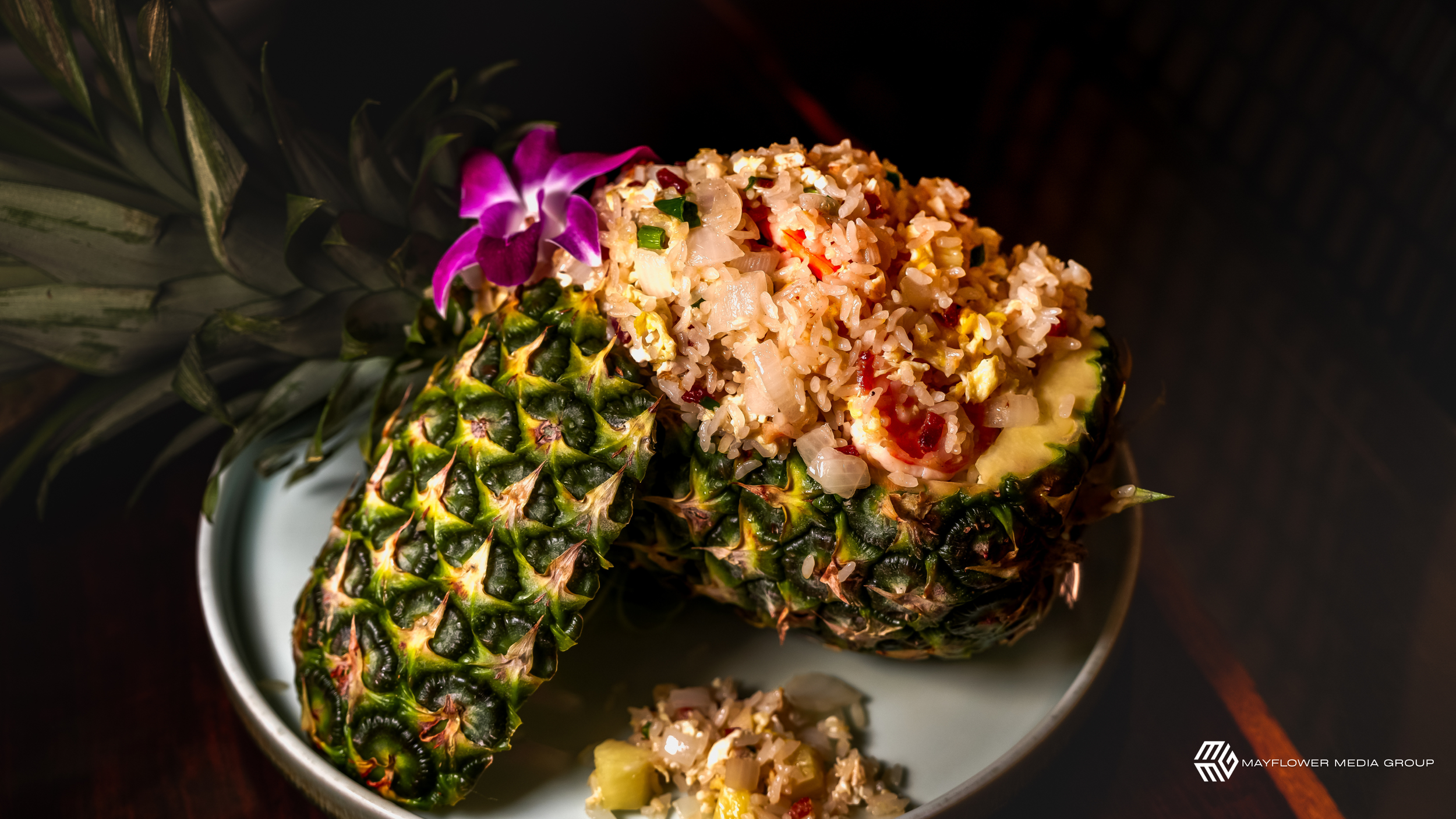 A hollowed pineapple filled with fried rice garnished with purple orchids on a white plate.