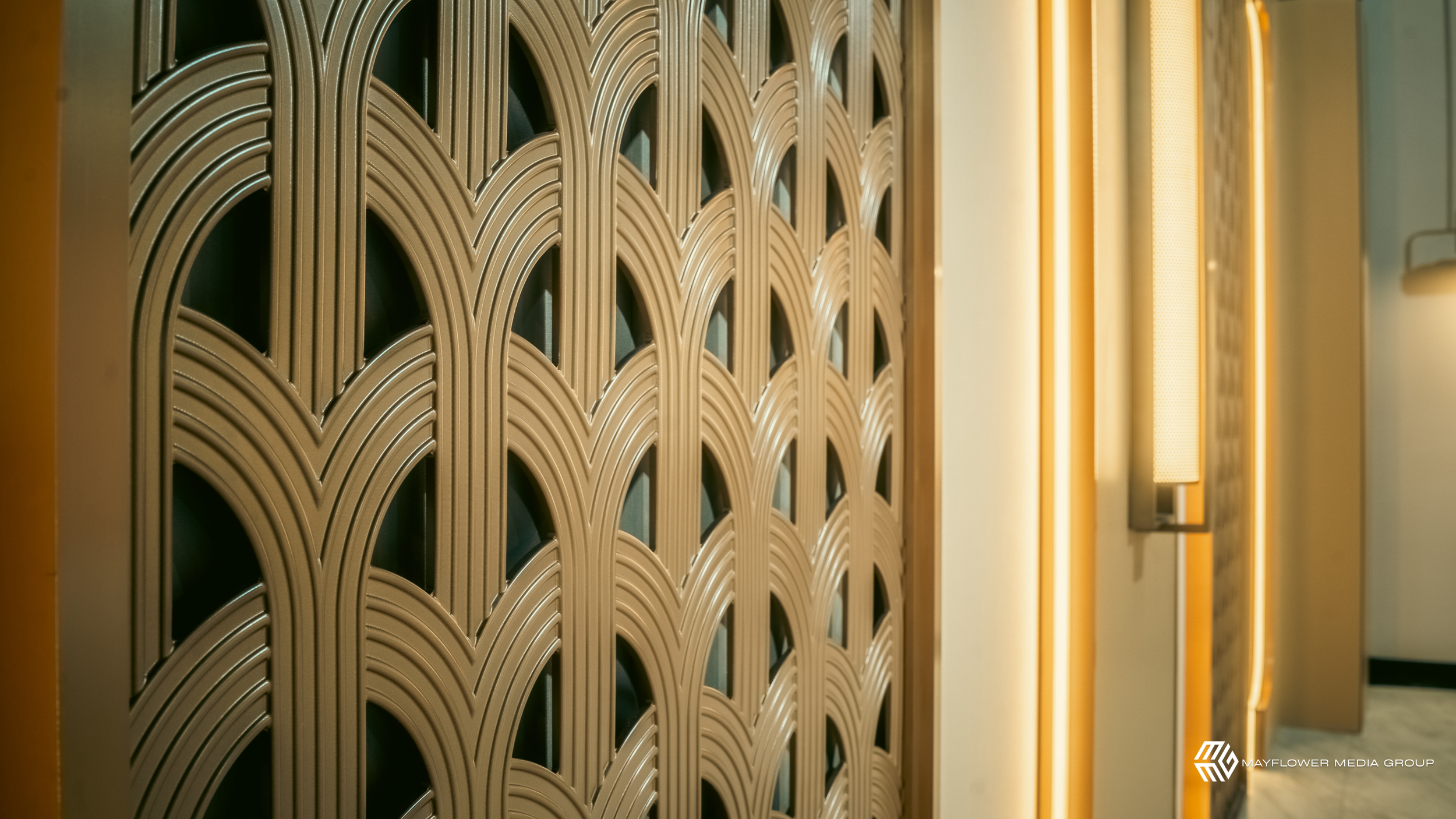 A decorative wall with a pattern of semi-circular arch shapes made of gold-colored material, illuminated by a vertical light fixture on the right side.