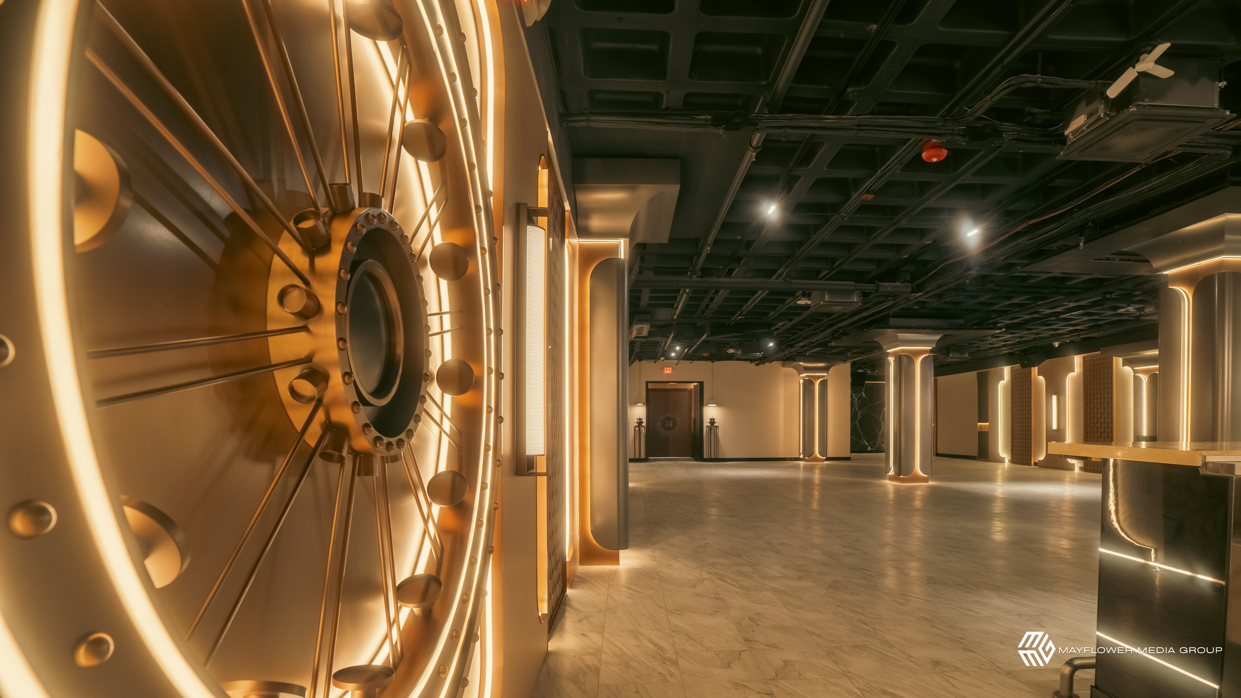 Photo of an elegant interior with a large, circular mechanical installation with lights on a wall, illuminated in warm tones, with a spacious hallway and modern architectural elements, including pillars with lighting accents.