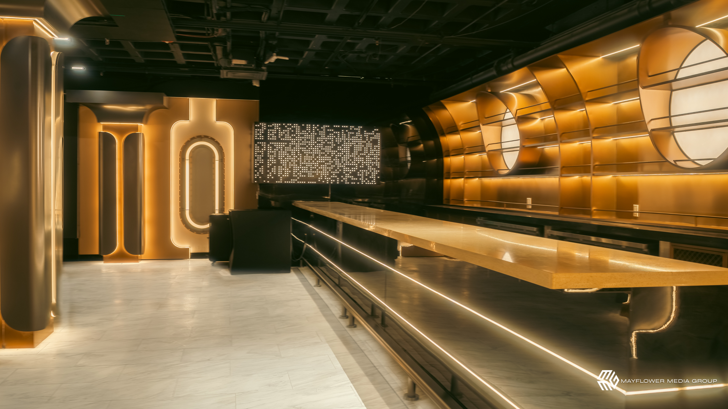 Interior of a stylish bar or restaurant with warm gold and black decor, illuminated wall features, a long marble or granite countertop, and modern lighting accents.