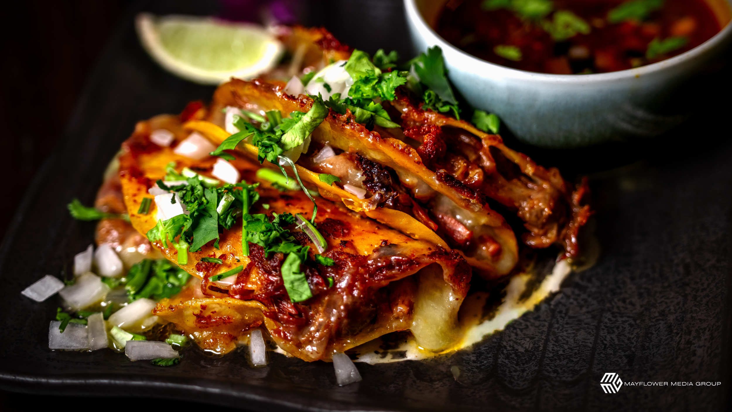 Close-up of Mexican food dish with melted cheese, chopped onions, and fresh cilantro, served on a black plate with a wedge of lime and a side of red sauce.