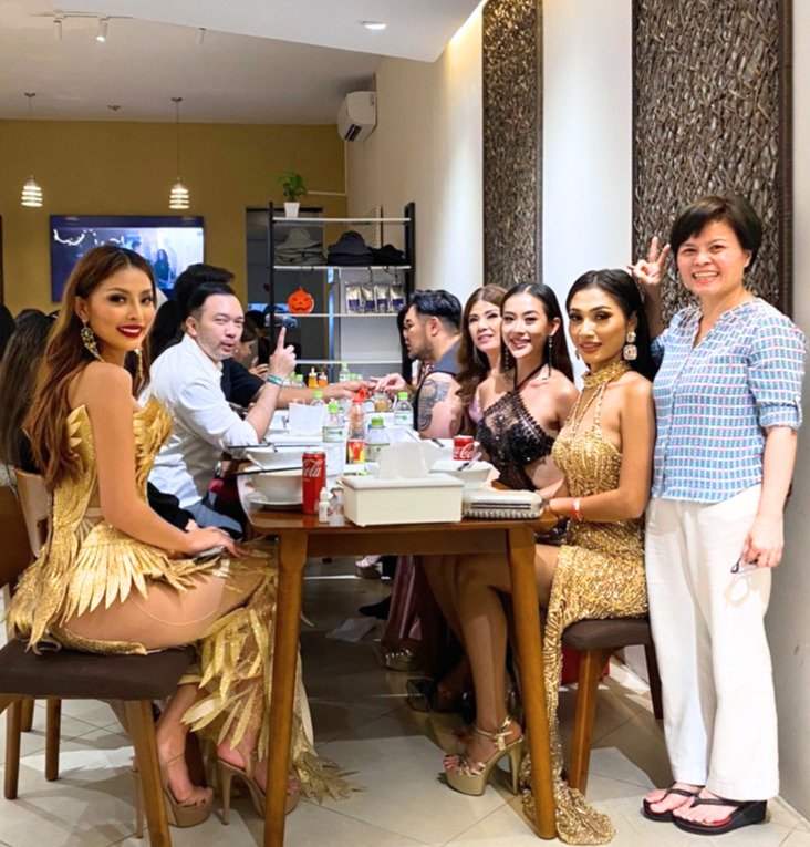 Group of women and one man sitting at a dining table in a restaurant, some women dressed in glamorous outfits, woman standing on the right making a peace sign.