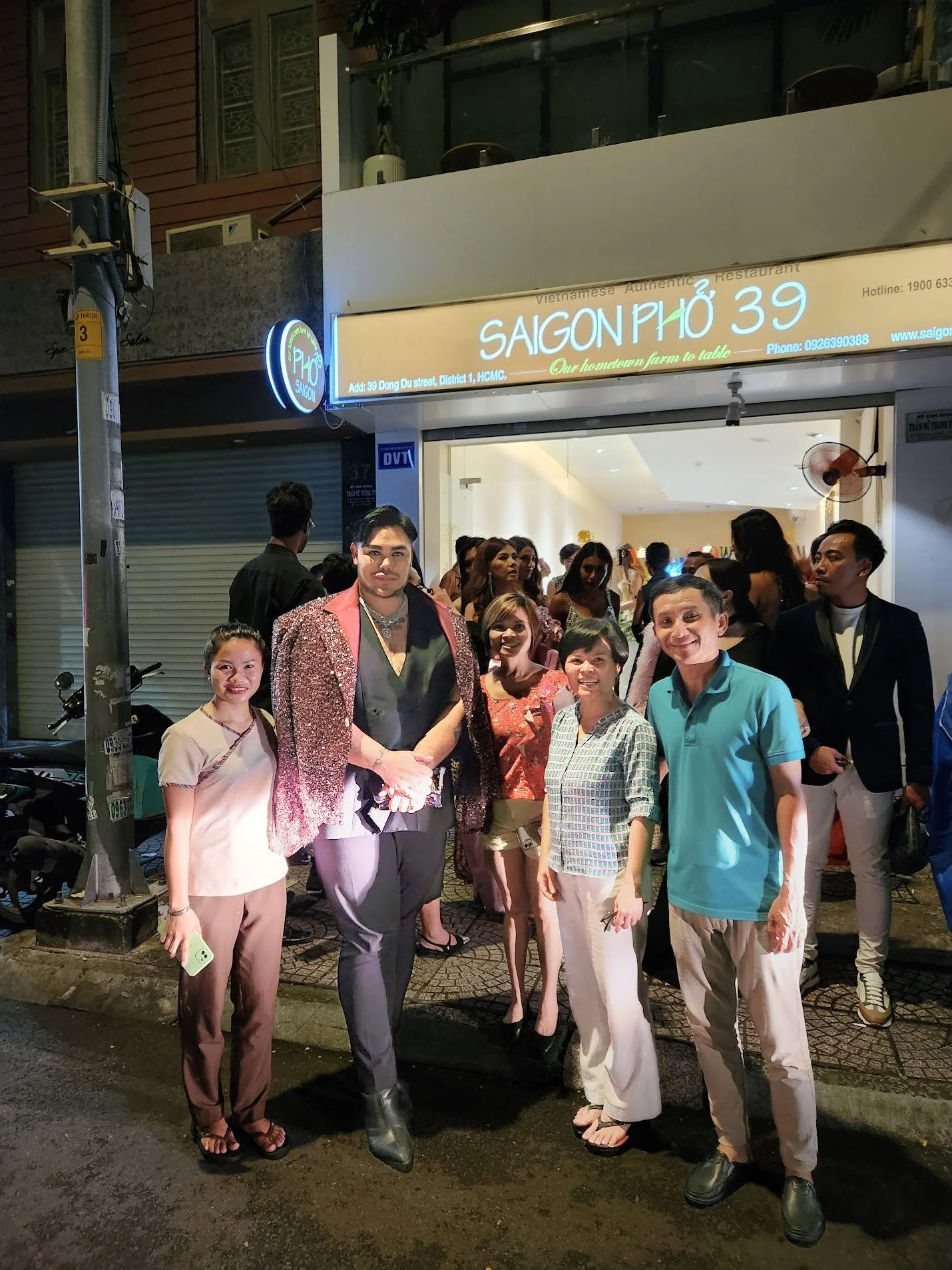 A group of people gathered outside a Vietnamese restaurant at night, with some individuals shaking hands, in front of a sign that reads 'Saigon Phở 39'.