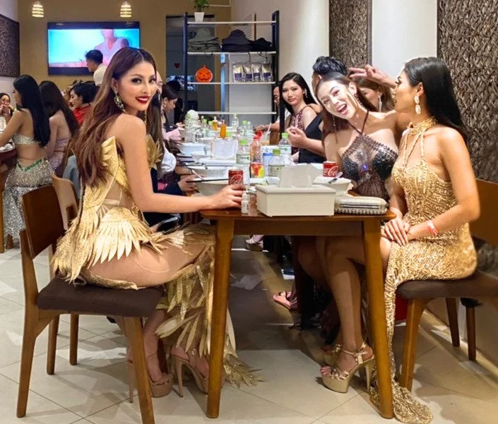 Group of women in glamorous dresses sitting around a table, enjoying a social gathering or celebration in a restaurant or event space.