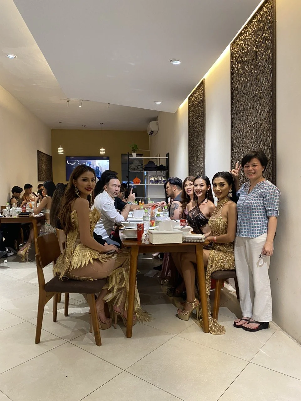 A group of people, including women in glamorous dresses, are sitting around a long table in a restaurant, celebrating an event.