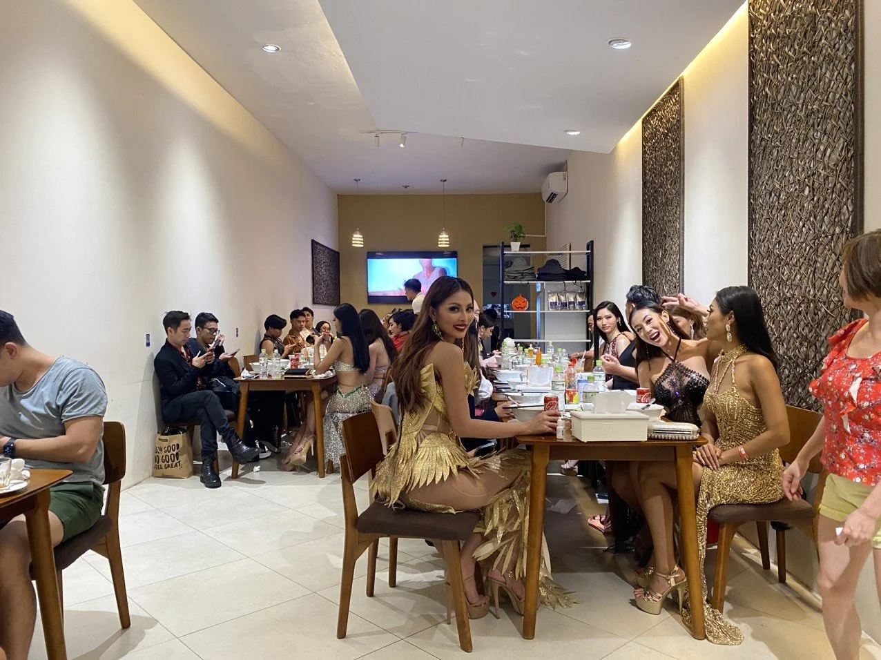 People sitting at a long table in a restaurant, some dressed in fancy attire, with a woman in a gold dress smiling at the camera, while others socialize and look at their phones.
