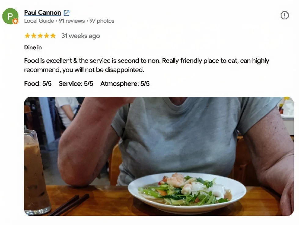 A person sitting at a restaurant table with a plate of stir-fried vegetables and shrimp in front of them, and a glass of iced coffee on the side.