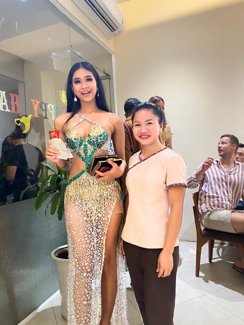 A woman in a colorful, ornate dress holding a drink and a clutch, standing next to another woman in a beige top and dark pants, in a room with people seated and talking, and 