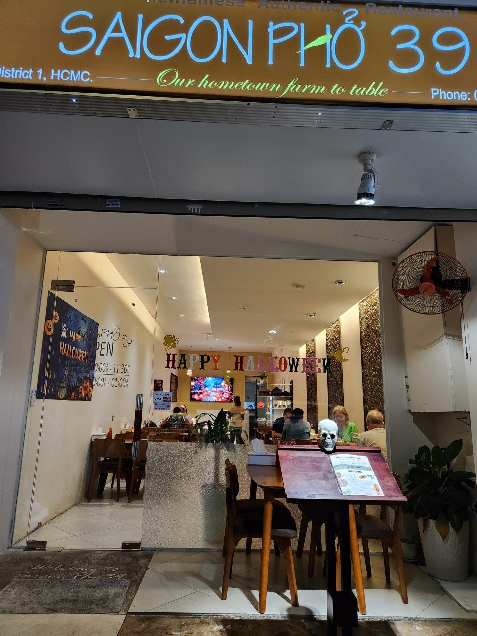 Entrance to Saigon Pho 39 Vietnamese restaurant decorated with Halloween banners, with seated customers inside and a TV screen visible.