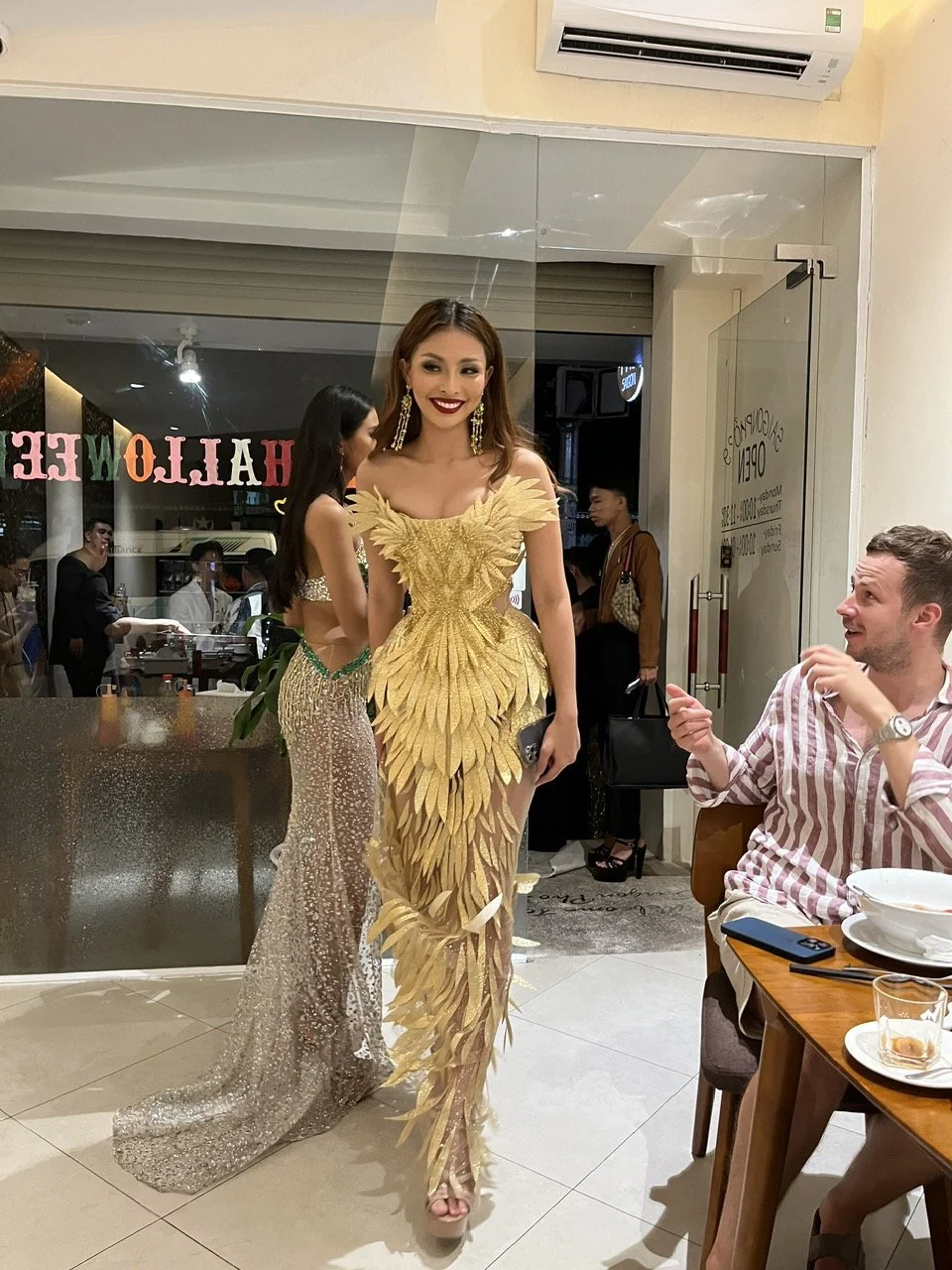 Two women in glamorous dresses, one in a gold feathered gown and the other in a sparkling silver gown, in a restaurant with a man at a table.