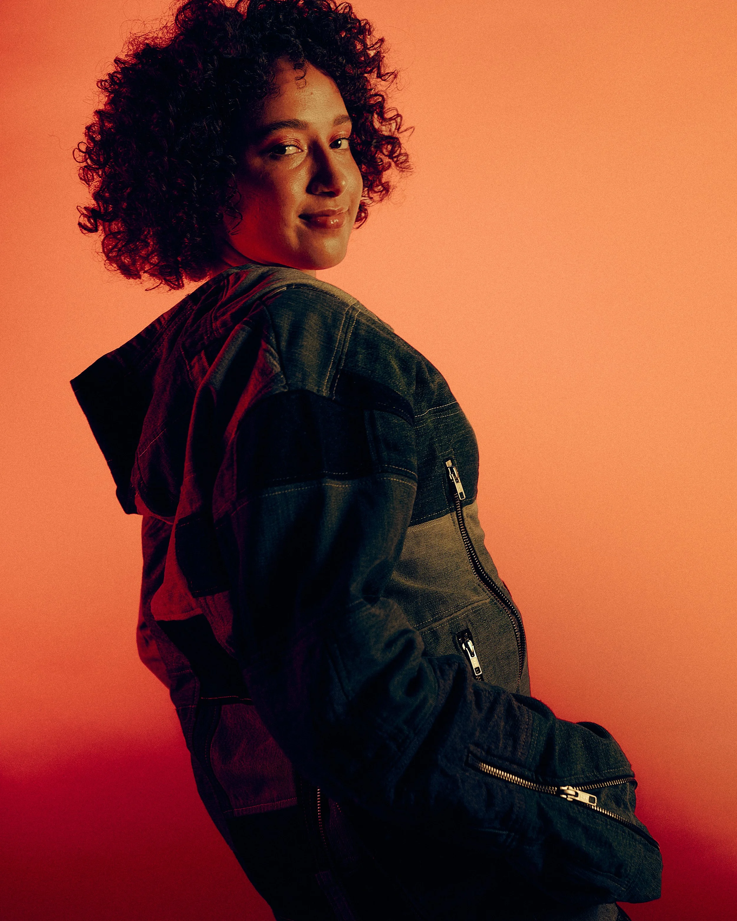 A woman with curly dark hair wearing a dark denim jacket, posing against a warm orange background.