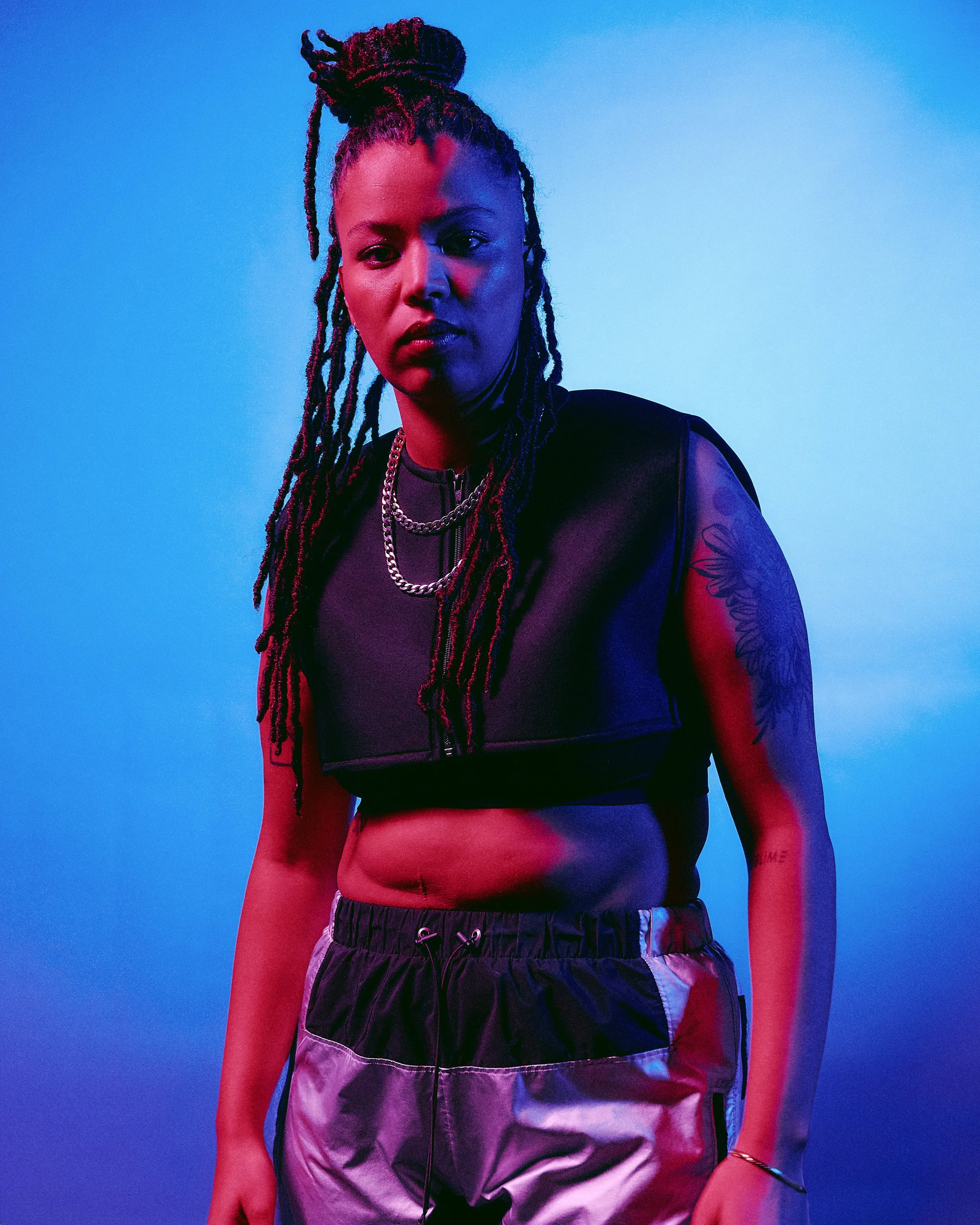 A woman with dreadlocks styled in an updo, wearing a black sleeveless top, layered chain necklaces, and patterned shorts, standing against a gradient blue background with colorful lighting.